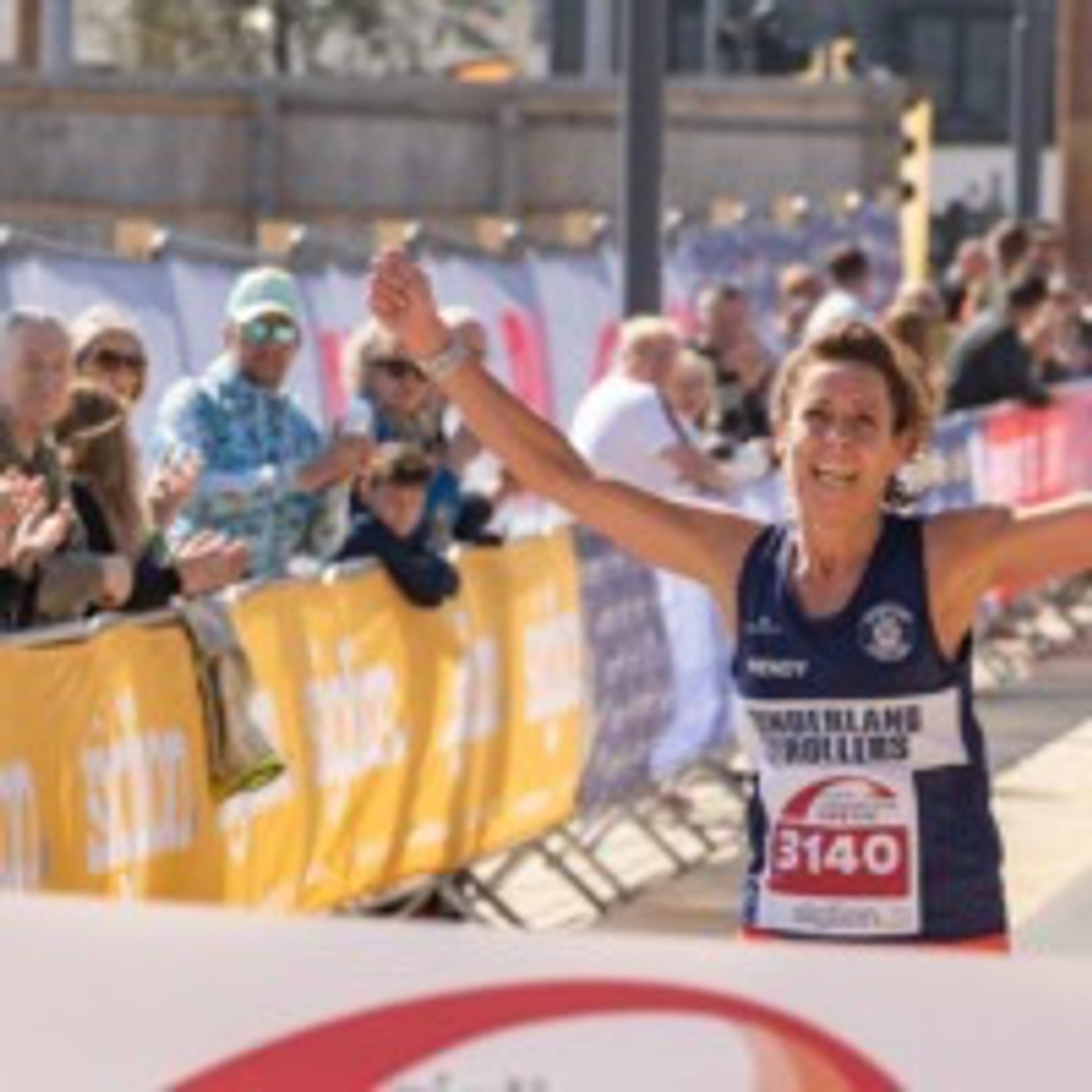 A runner wearing a dark blue sleeveless shirt with race bib number 3140 crosses the finish line with arms raised in celebration. Spectators cheer from behind yellow barricades. The sun shines brightly, contributing to a festive atmosphere at the event.