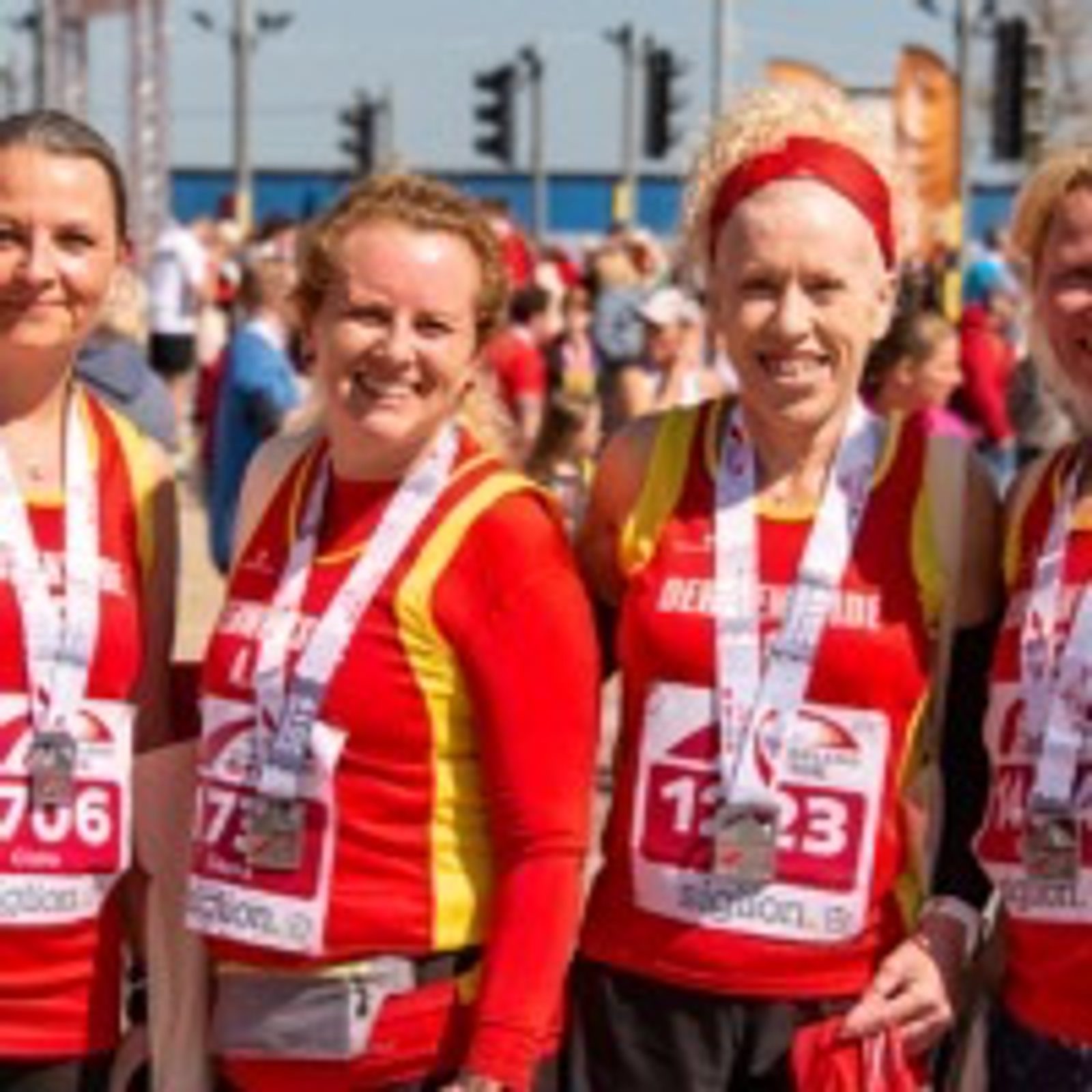 Five individuals wearing red and yellow athletic attire stand together with medals around their necks, smiling at the camera. They are outdoors, and other people in the background are also dressed in athletic clothing, suggesting a race or athletic event.