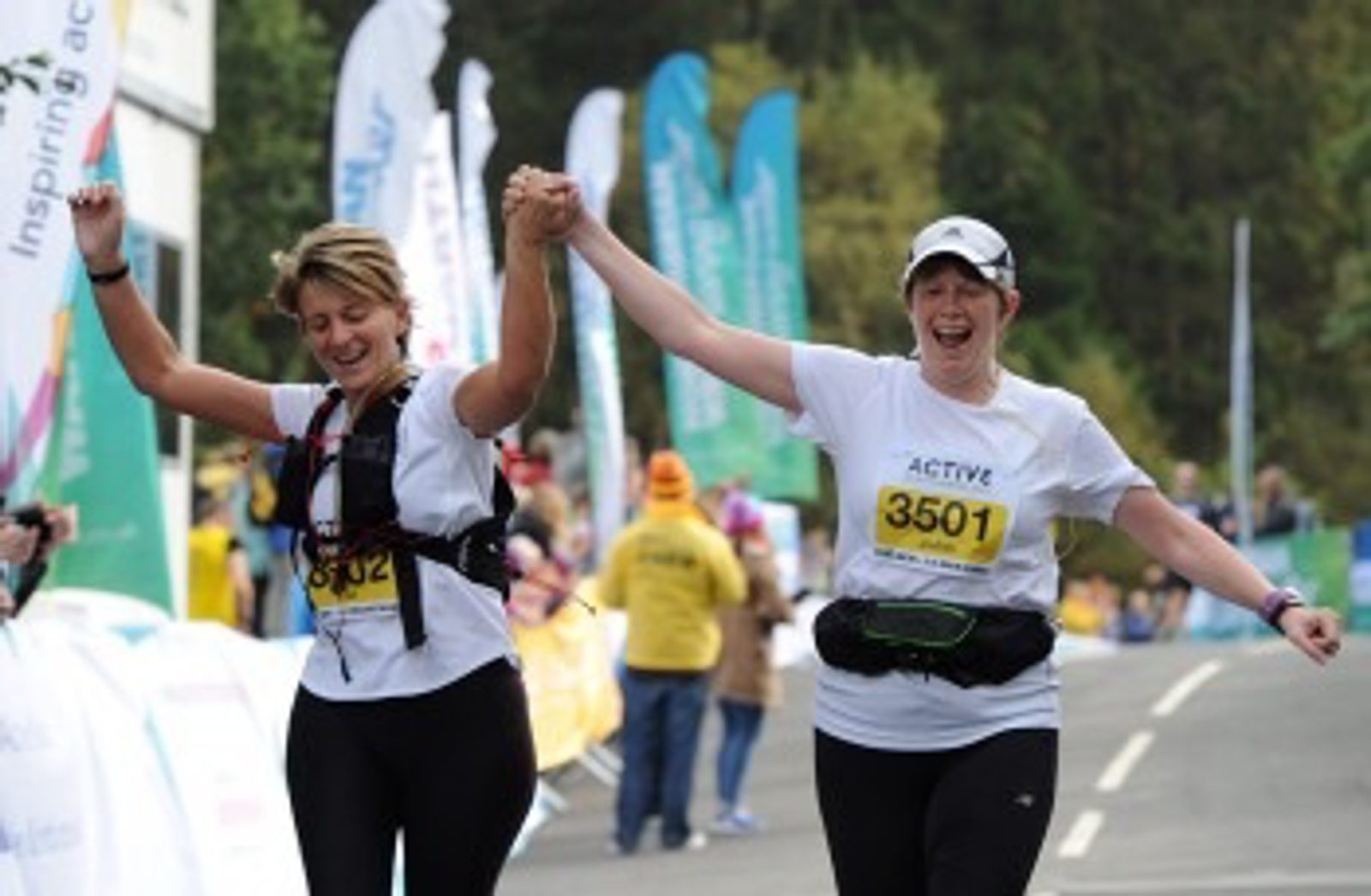 Two women in athletic gear hold hands and raise them victoriously as they cross a race finish line. Both are smiling and appear happy. They are wearing white shirts with bib numbers 3501 and 3502. Spectators and colorful flags are visible in the background.