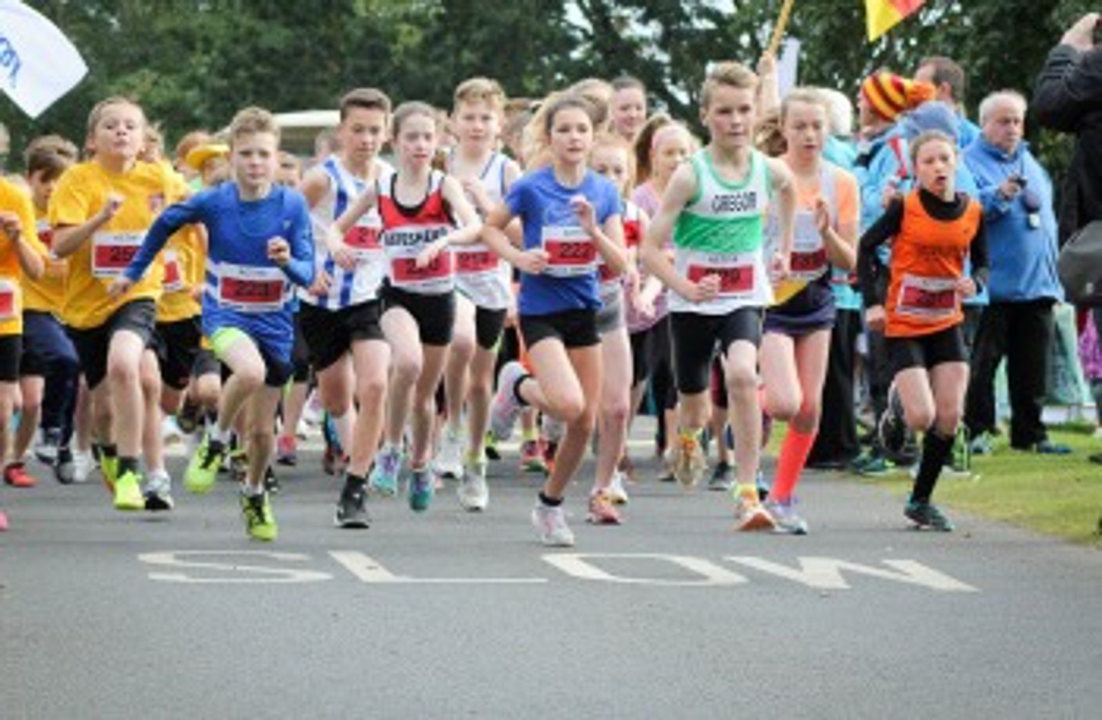 A group of young runners dressed in colorful athletic wear are starting a race on a road marked with the word "SLOW." Spectators and participants in yellow shirts stand in the background, and trees line the course on a clear day.