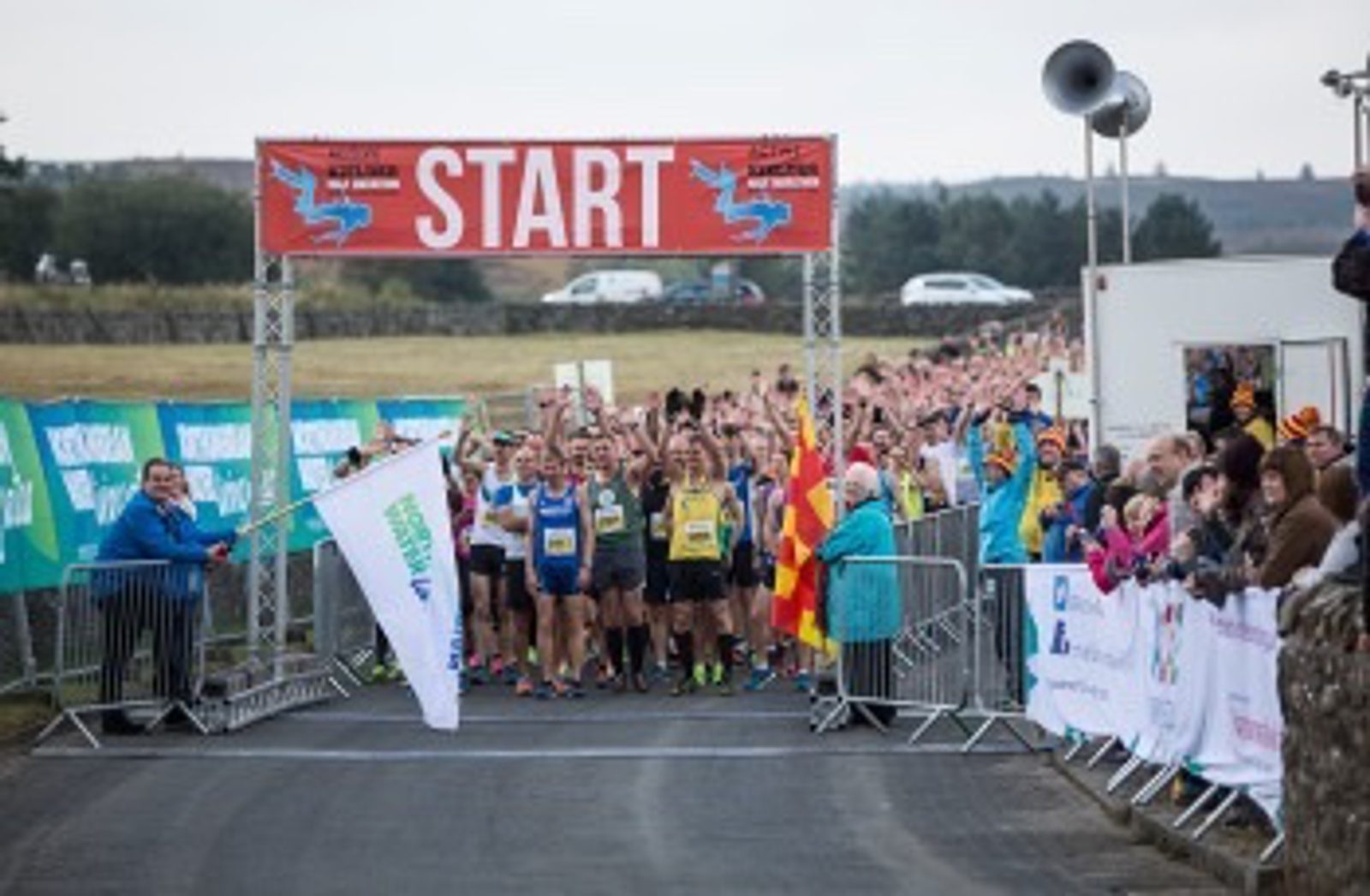 A large group of runners gathers at a starting line for a race, beneath a banner reading "START." Spectators cheer from the sides, and officials in colorful jackets prepare to begin the event. The background features open countryside and parked cars.
