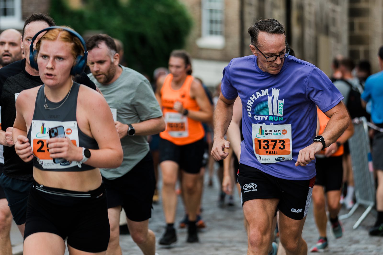 A group of runners participating in a city race, with numbered bibs pinned to their shirts. The focus is on a woman in black gear and a man in a purple shirt and glasses, both running on a cobblestone street.