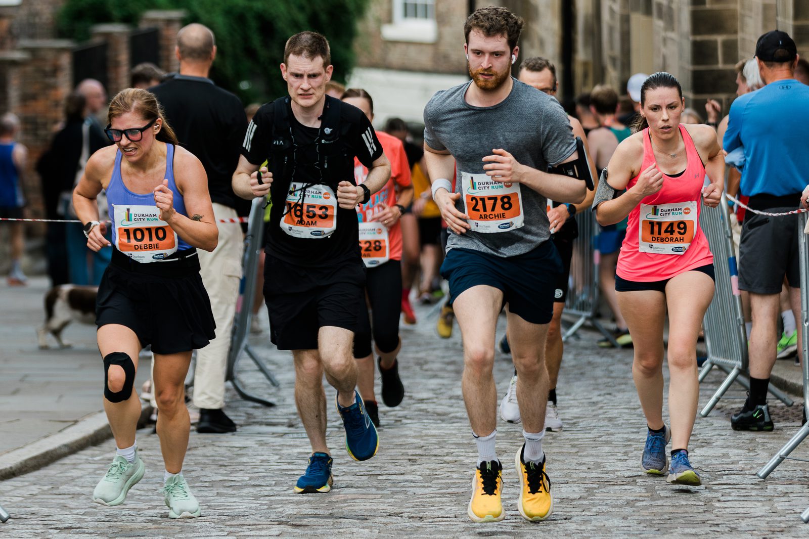 Four runners in athletic gear, each wearing numbered race bibs, are competing in a road race on a cobblestone street. Spectators and other participants are visible in the background.