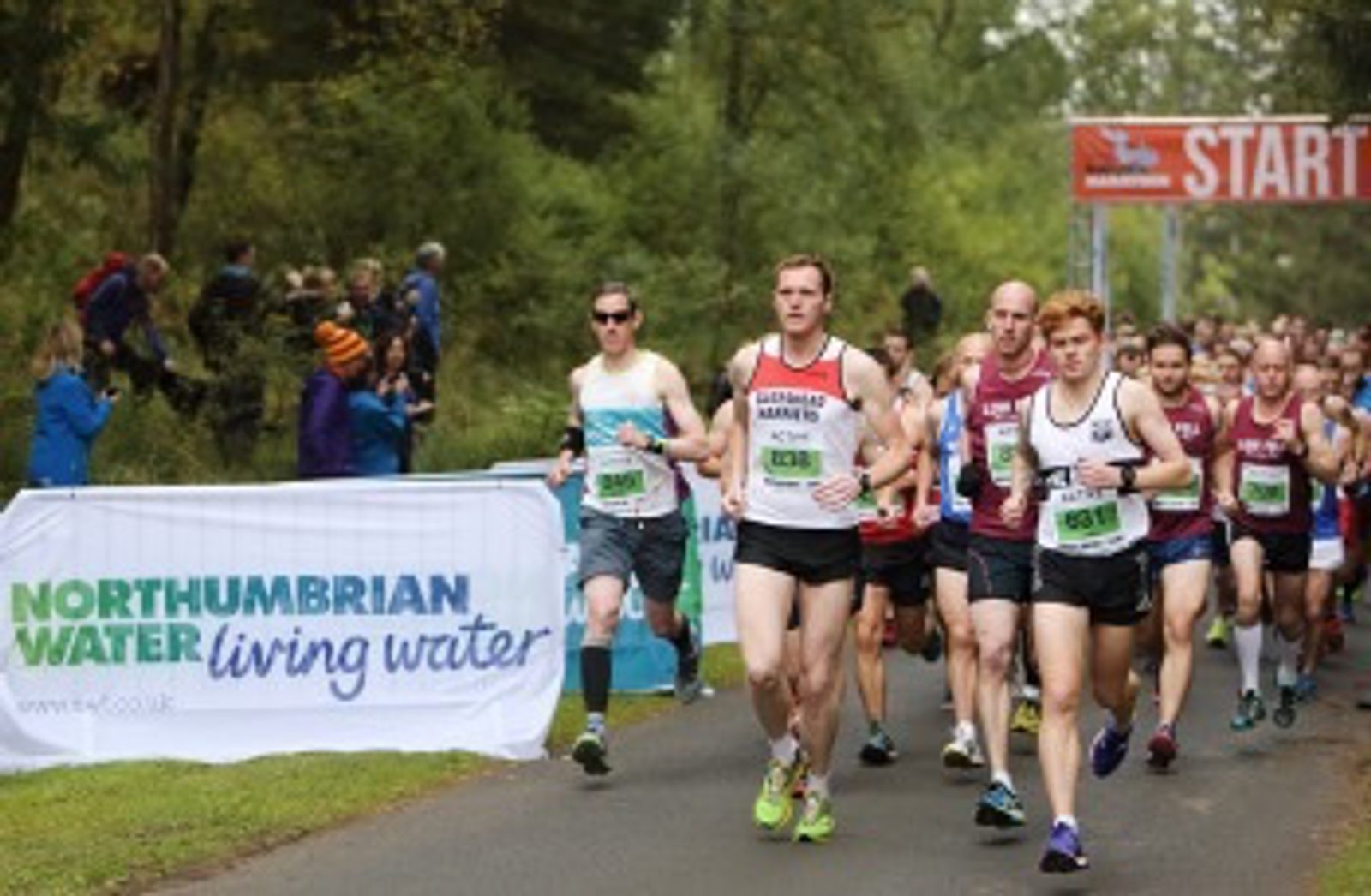 A group of runners are participating in a race on a paved road through a forested area. The runners are wearing different athletic outfits and bib numbers. A banner reading "Northumbrian Water living water" is displayed on the left. A "Start" banner is visible in the background.