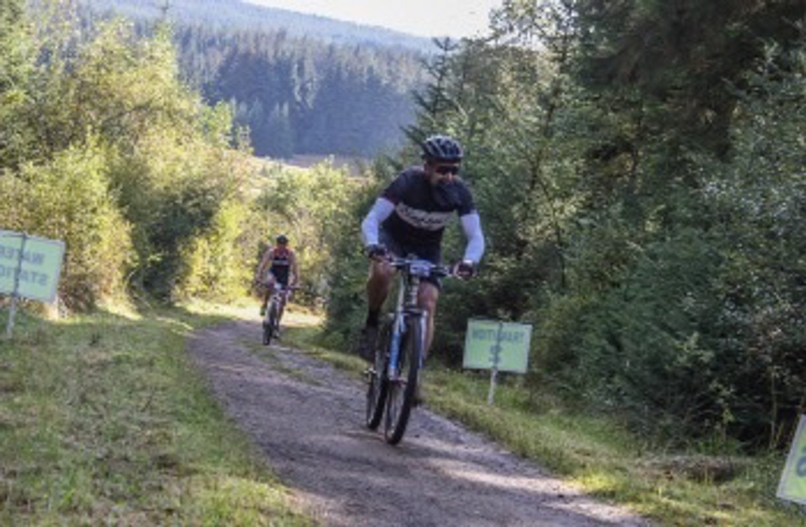 Two cyclists ride along a forest trail with greenery and trees surrounding them. One cyclist leads in the foreground wearing a helmet and sunglasses, while the other follows in the distance. The path is dirt, and signs can be seen along the sides of the trail.