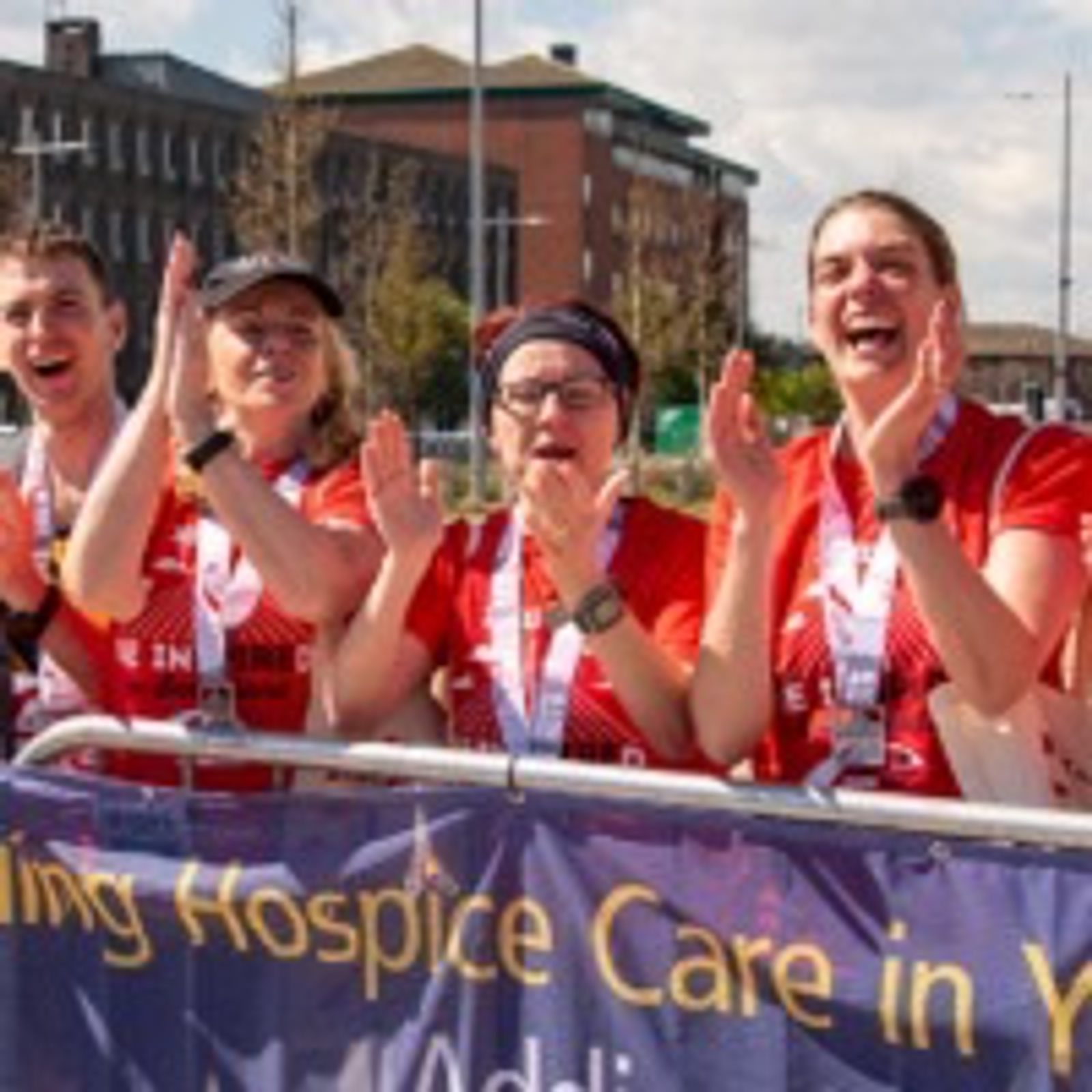 A group of four people wearing sports attire and medals enthusiastically clap and smile behind a banner that partially reads, "Hospice Care." The background shows a brick building, trees, and a street lamp under a sunny sky.