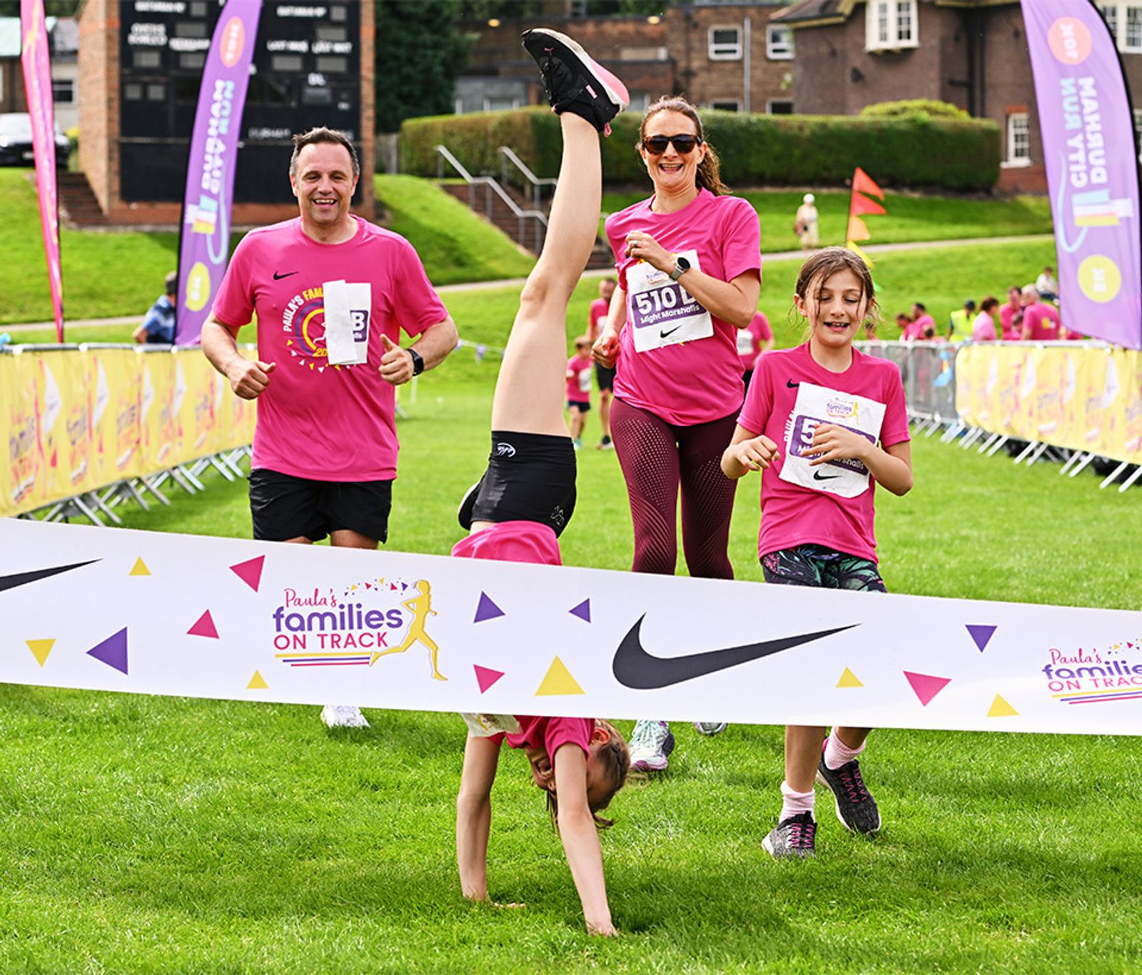 A family in pink shirts finishes a race. The young girl leads with a cartwheel across the finish line, while the parents and another child follow, smiling. Colorful banners and cheering spectators line the grass course.