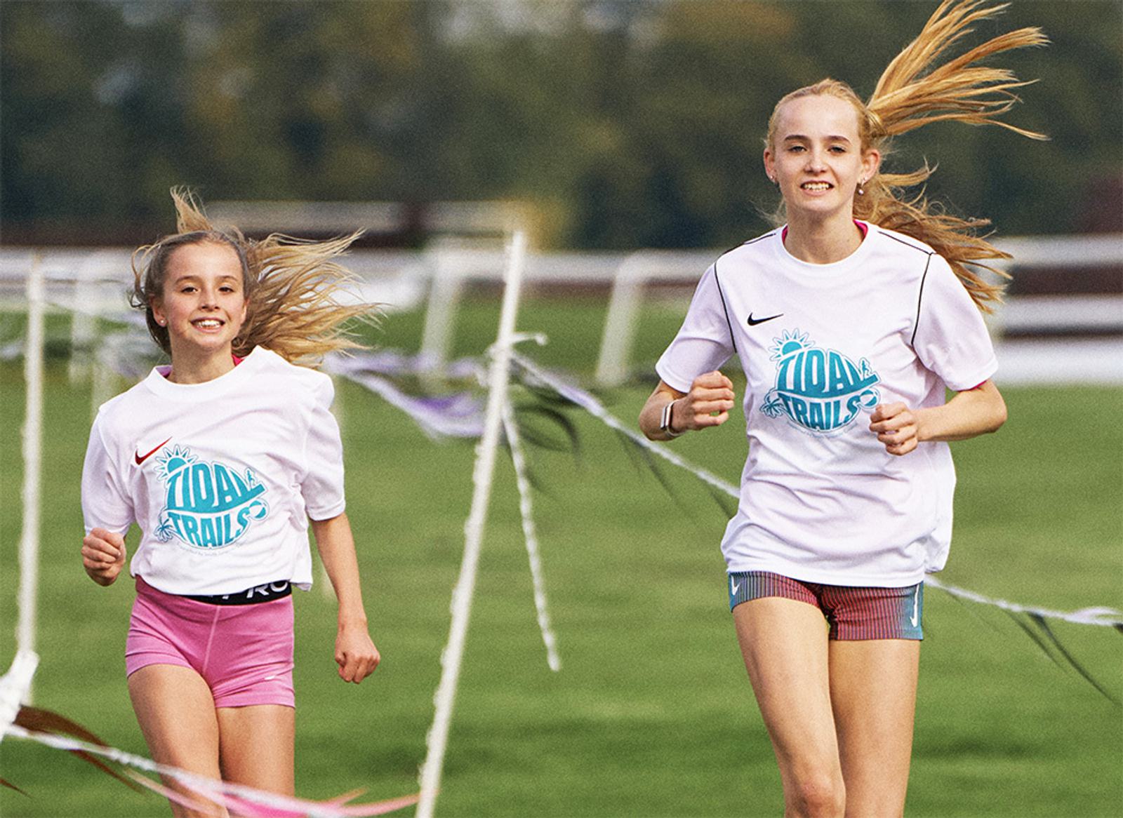 Two young girls running together on a grassy path, wearing matching white T-shirts with "IDOL TRAILS" written on them. They both have long blonde hair tied back, and the background shows a blurred view of trees and a fence.