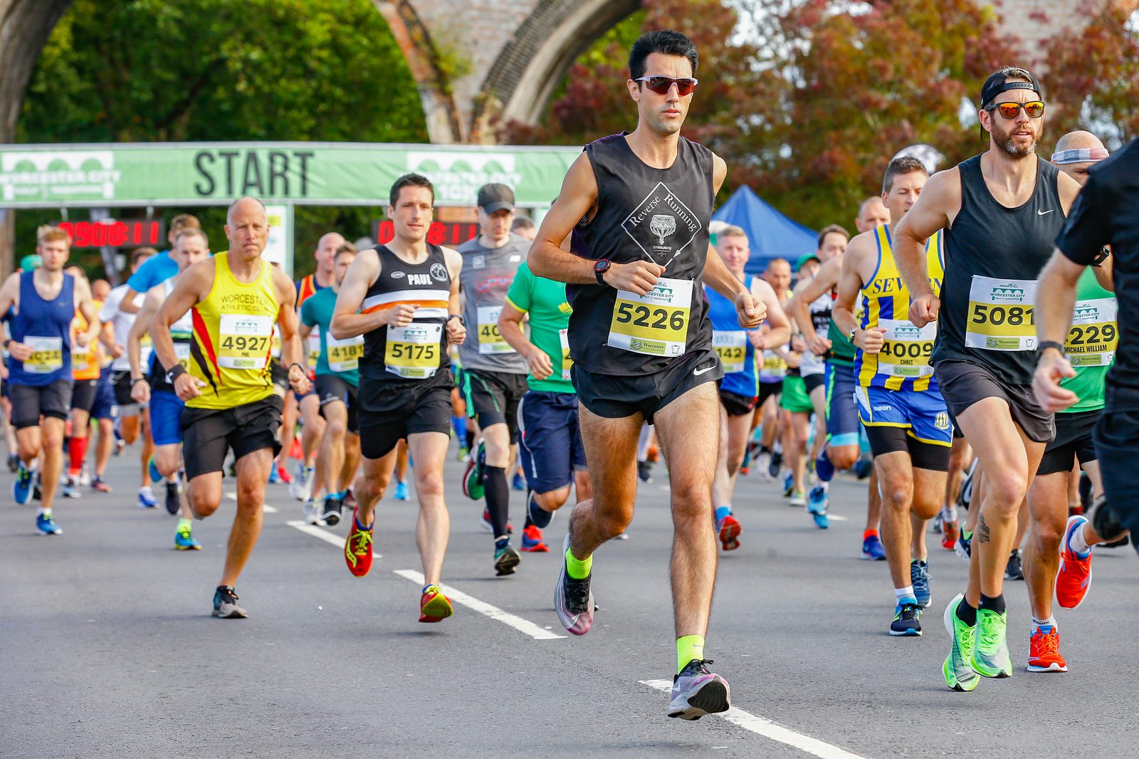 A group of runners, both men, and women, are participating in a race. Some are wearing colorful athletic gear, sunglasses, and race bibs with numbers. They are running on a paved road under a green banner that says "START." Trees and an archway are visible in the background.
