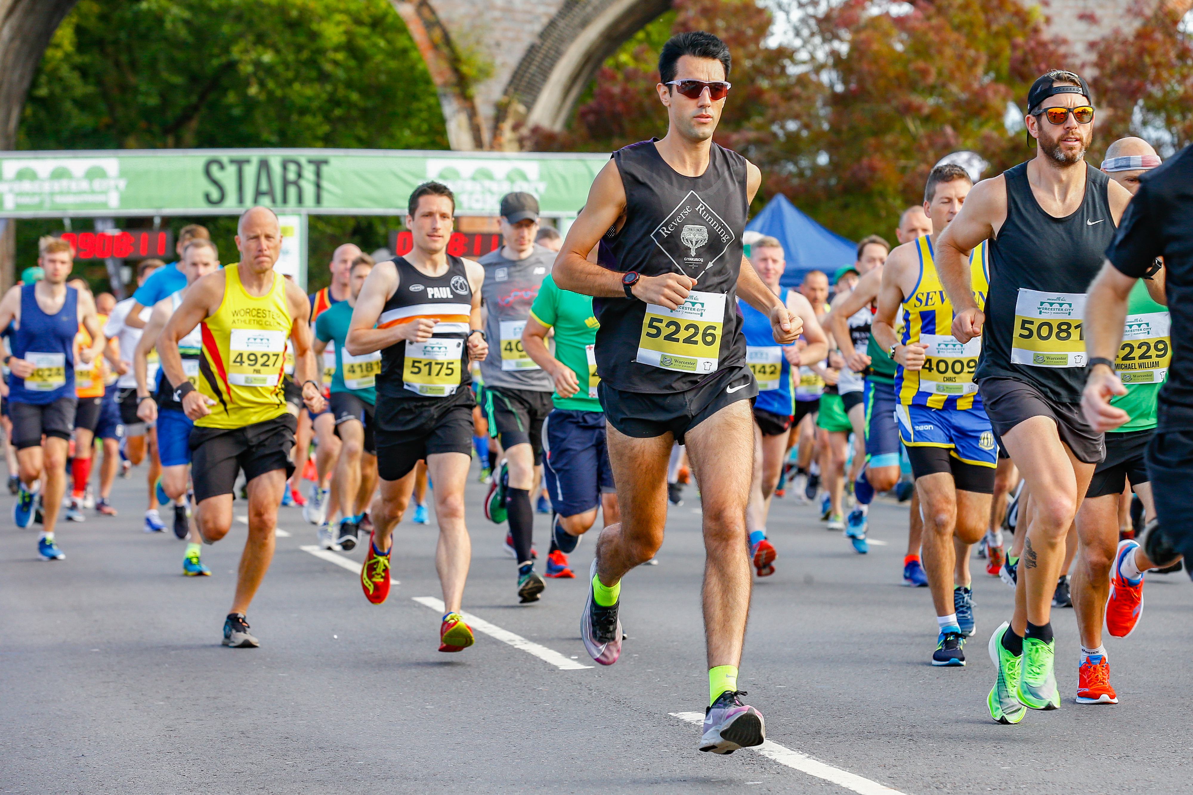 A group of runners, both men, and women, are participating in a race. Some are wearing colorful athletic gear, sunglasses, and race bibs with numbers. They are running on a paved road under a green banner that says "START." Trees and an archway are visible in the background.