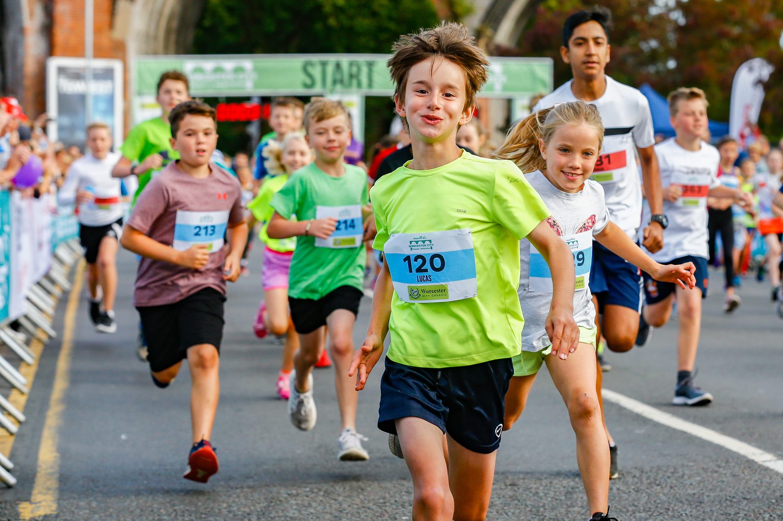 A group of children are running in a race, all wearing race bibs with numbers. The boy in the foreground, wearing a lime green shirt and black shorts, appears joyful. Other children, boys and girls, also look focused and energetic as they run on the road.