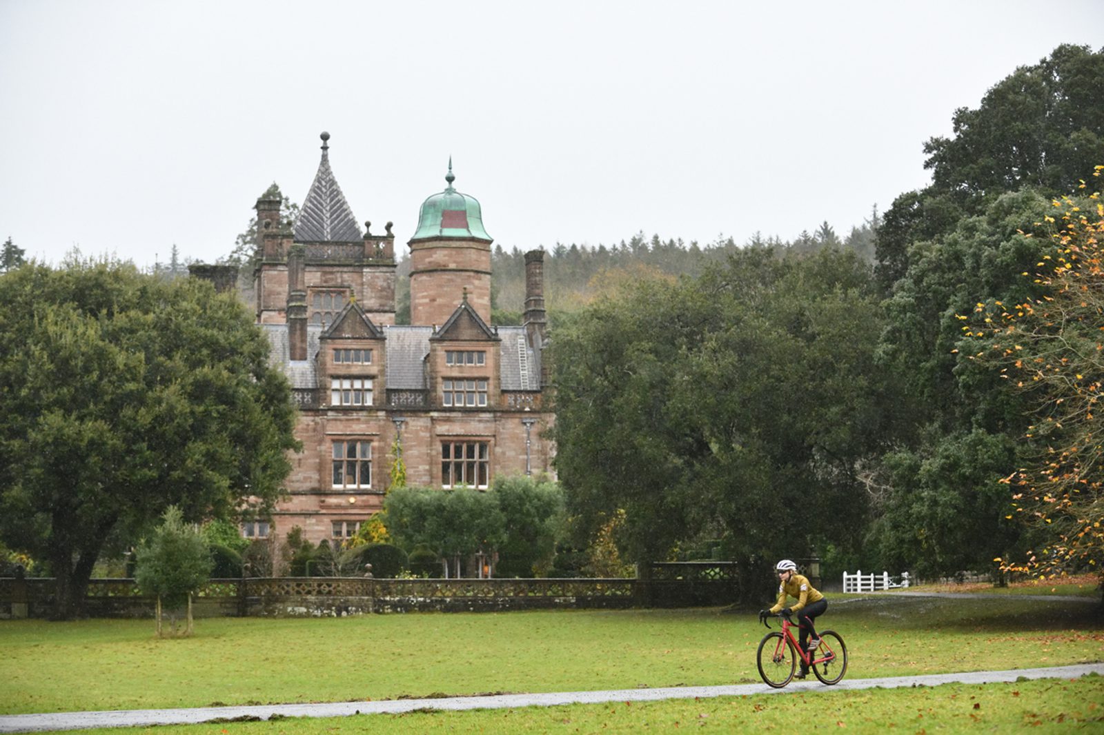 A cyclist in a yellow jacket rides a red bicycle on a paved path through a grassy park, with a large, historic castle featuring towers and a green dome visible in the background surrounded by trees. The sky is overcast.