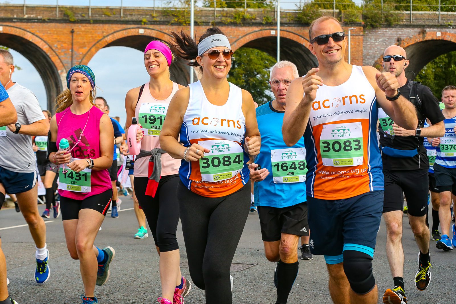 A group of runners participating in a race. Two center runners wearing 'Acorns' shirts are smiling and giving thumbs up. They are surrounded by other runners wearing bib numbers, some in bright athletic clothing. A brick viaduct and spectators are visible in the background.