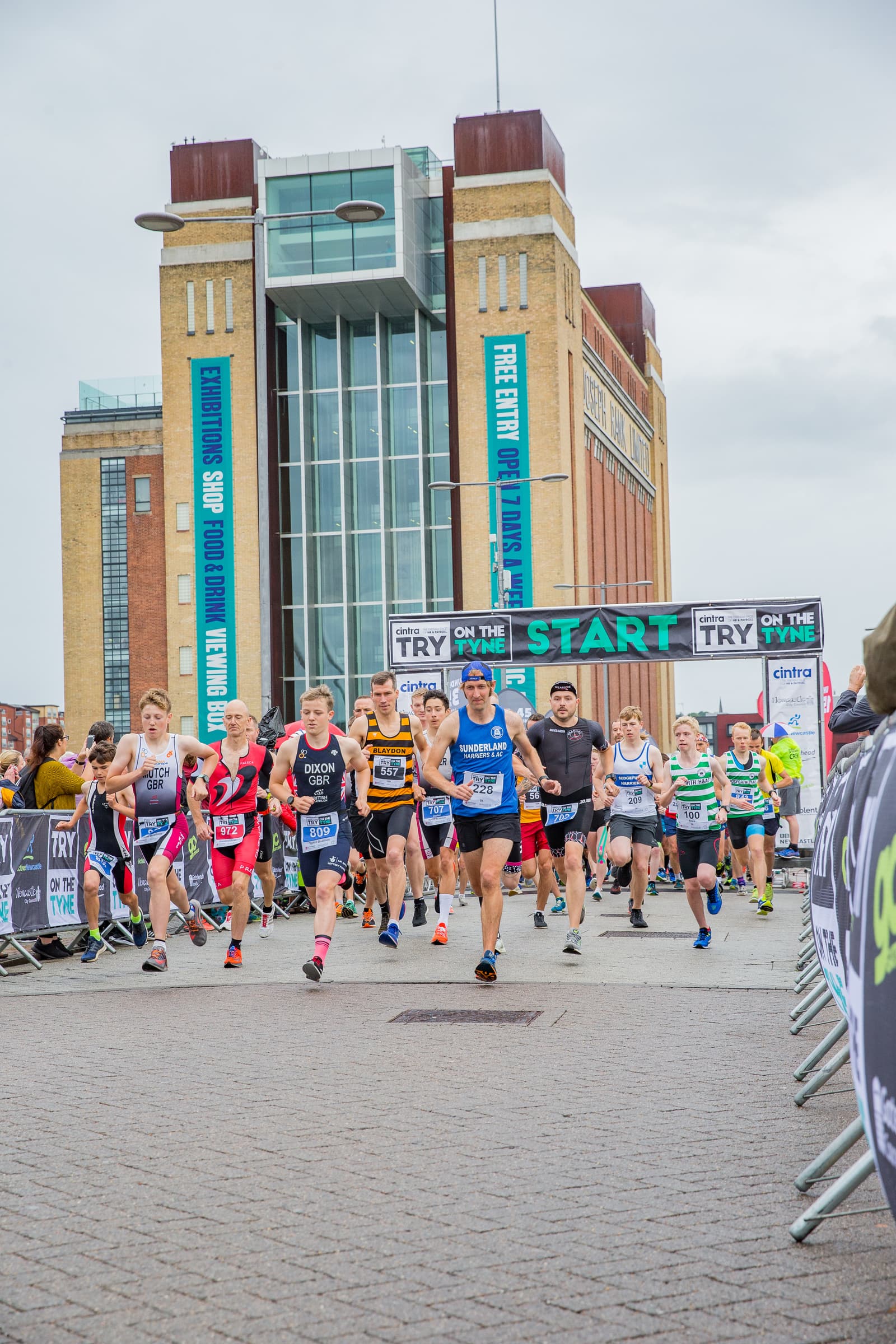 A group of runners, wearing colorful athletic gear and numbered bibs, sprint from the starting line of a race. The race course is lined with banners and spectators. In the background, a tall, brick building with large windows and banners is visible.