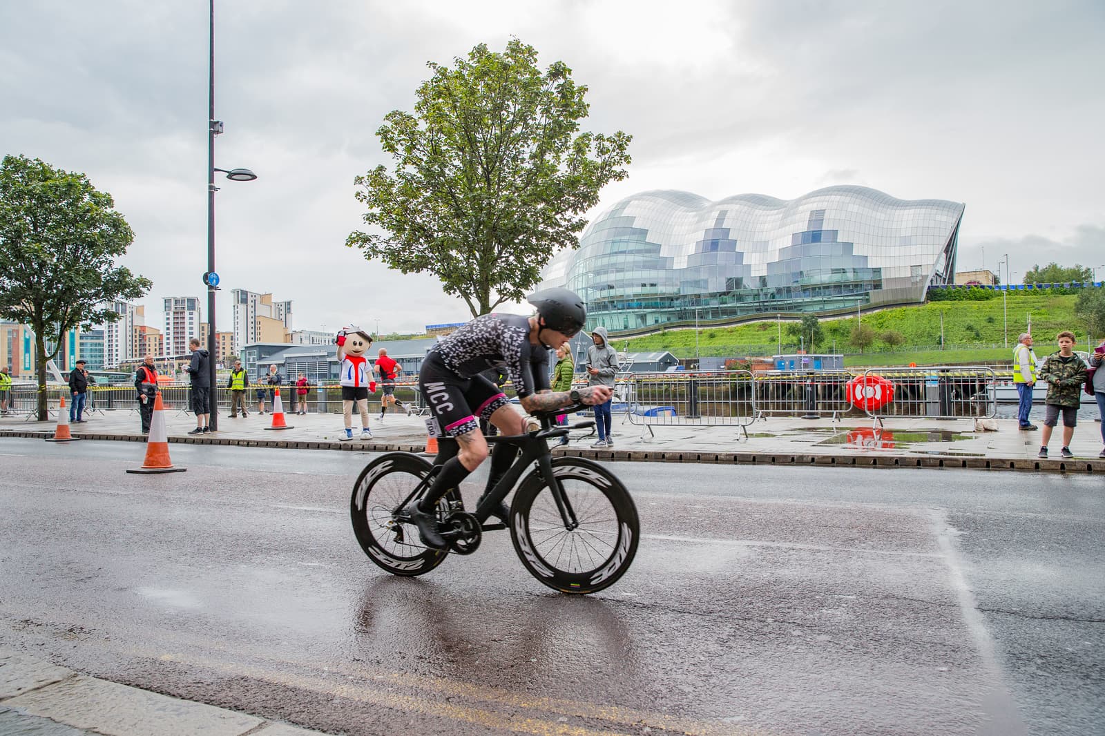 A cyclist in a race rides on a wet road with an aerodynamic helmet and a specialized bike. Spectators and event staff watch from the sidewalks. The background features modern architecture, trees, and traffic cones outlining the route. The sky is overcast.