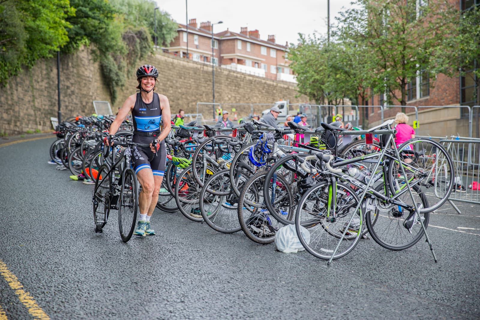 A person in athletic gear and a helmet walks while holding a bicycle. They are amongst a row of parked bicycles on the side of a street, with buildings and trees in the background. The scene appears to be at a cycling or triathlon event.