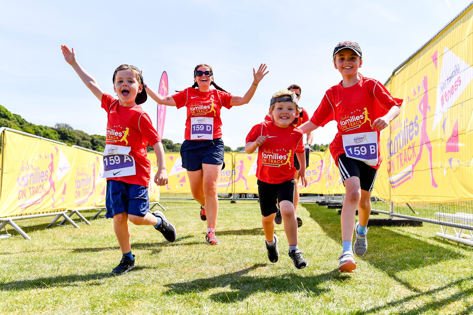A group of three children and one adult, all wearing matching red shirts, enthusiastically cross a finish line during an outdoor family race event. They are smiling and cheering, with arms raised in celebration. Bright yellow banners line the pathway.