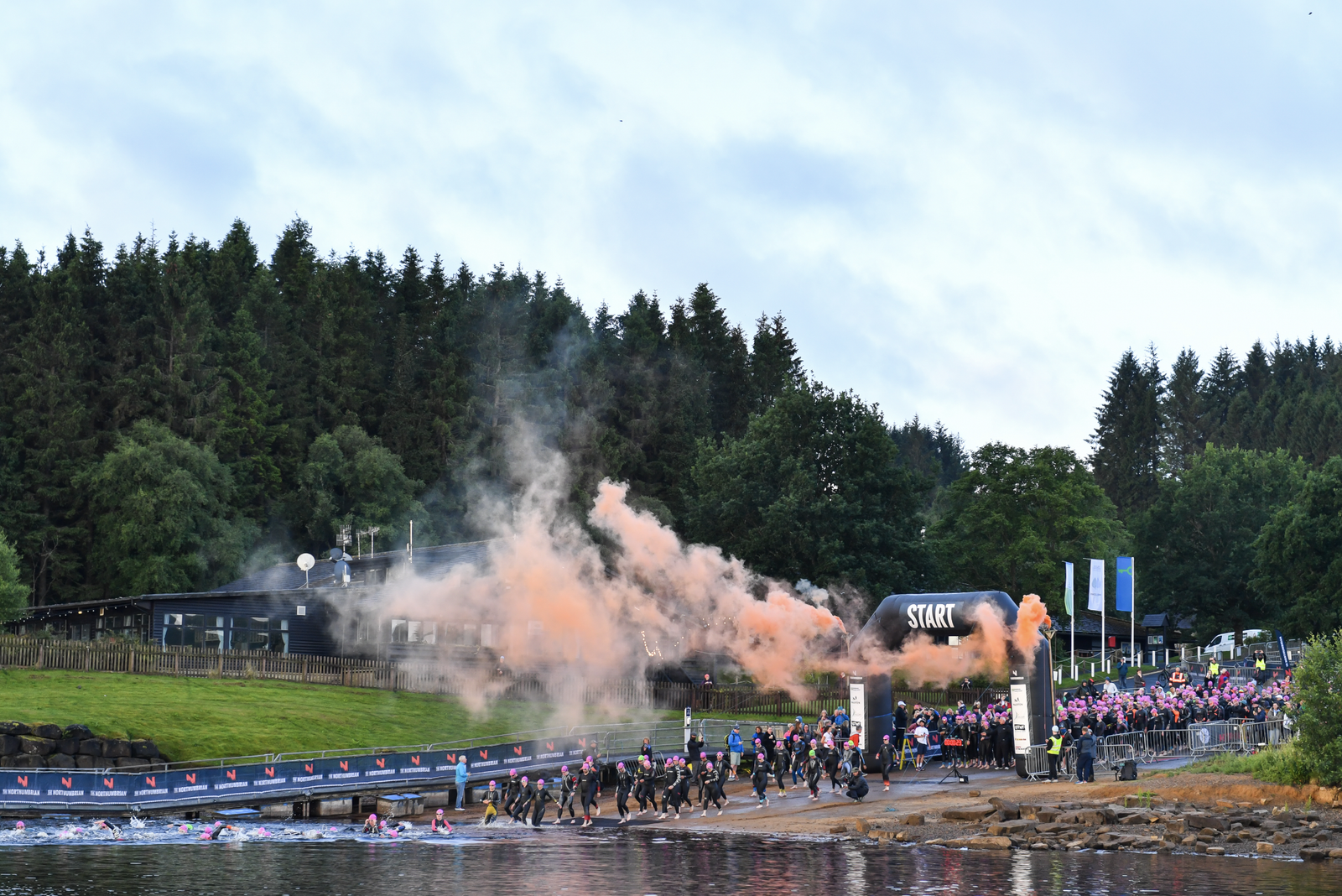 A triathlon starts with participants running into the water under a starting arch that emits orange smoke. A crowd watches by the side, and trees form the backdrop. The sky is overcast, and the scene is filled with anticipation and energy.