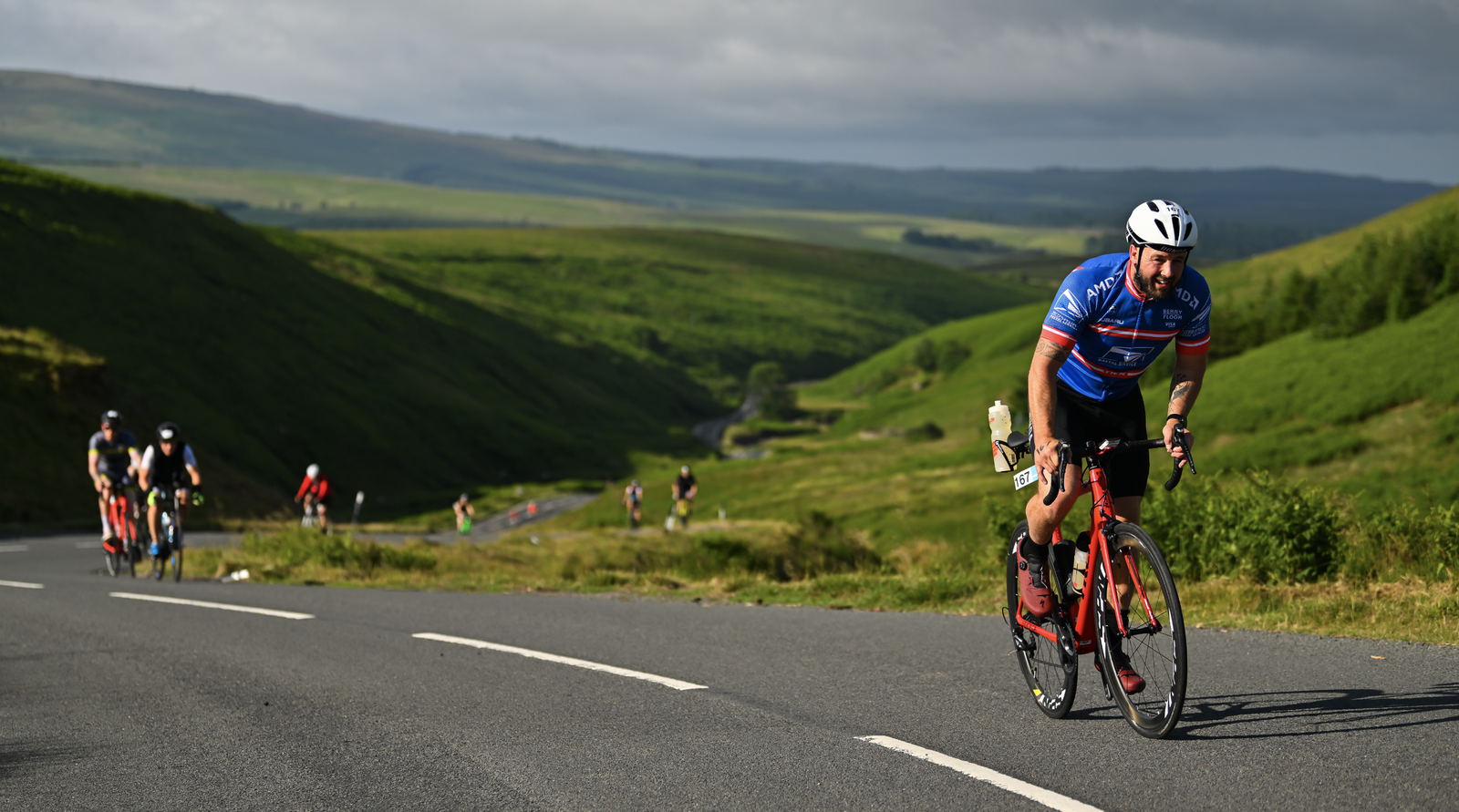 A cyclist wearing a blue and red jersey and white helmet rides uphill on a winding road surrounded by rolling green hills. Other cyclists are seen further back along the road. The sky is cloudy, casting shadows on the landscape.