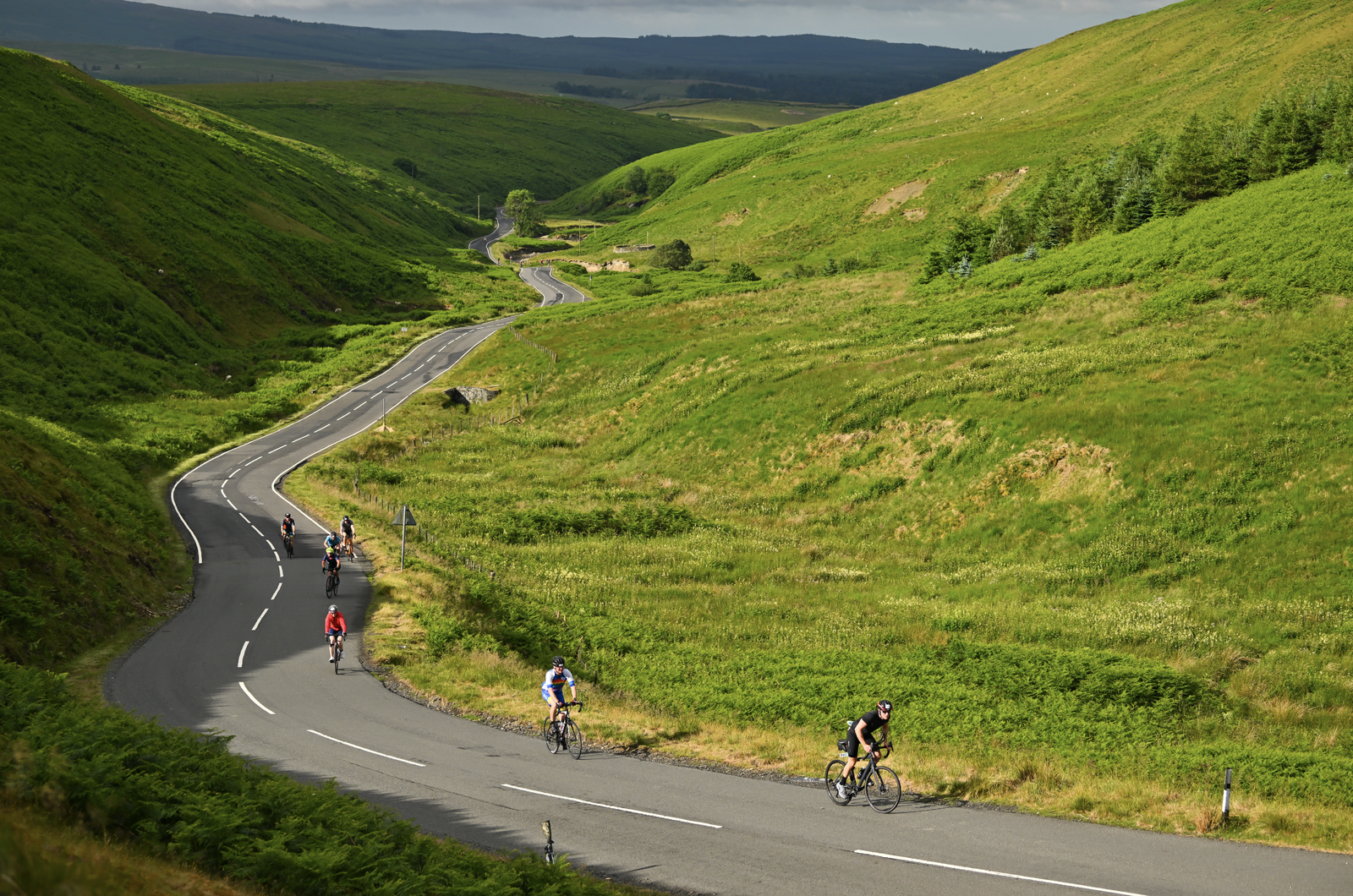 A scenic view of a winding road through lush green hills with several cyclists riding uphill. The landscape features rolling hills and dense greenery under a partly cloudy sky. The cyclists are spread out on the road, pedaling at different paces.