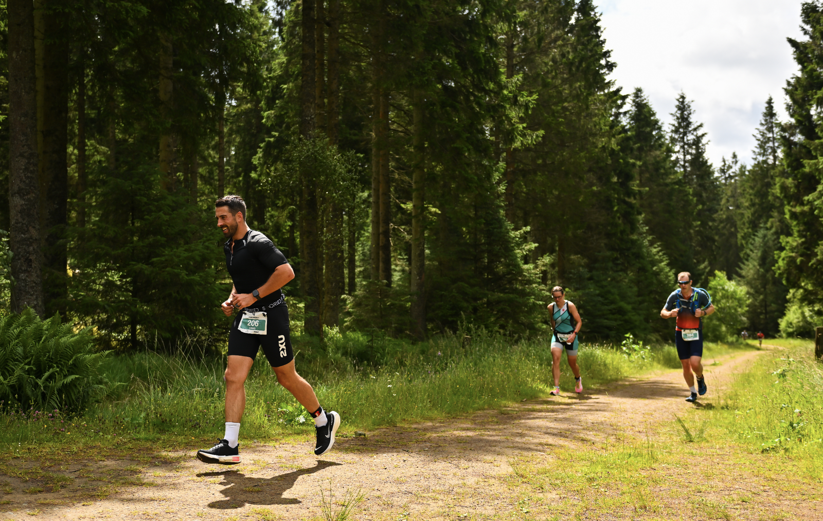 Three athletes run along a dirt trail in a lush, green forest. The runner in the foreground is wearing black sportswear, while the two runners in the background wear lighter, colorful gear. The sunlight filters through the trees, casting dappled shadows on the path.