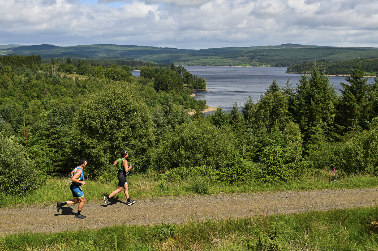 Two people in athletic gear run on a gravel path through a lush, green forest with a large lake and rolling hills in the background. The sky is partly cloudy, casting dappled sunlight over the serene landscape.