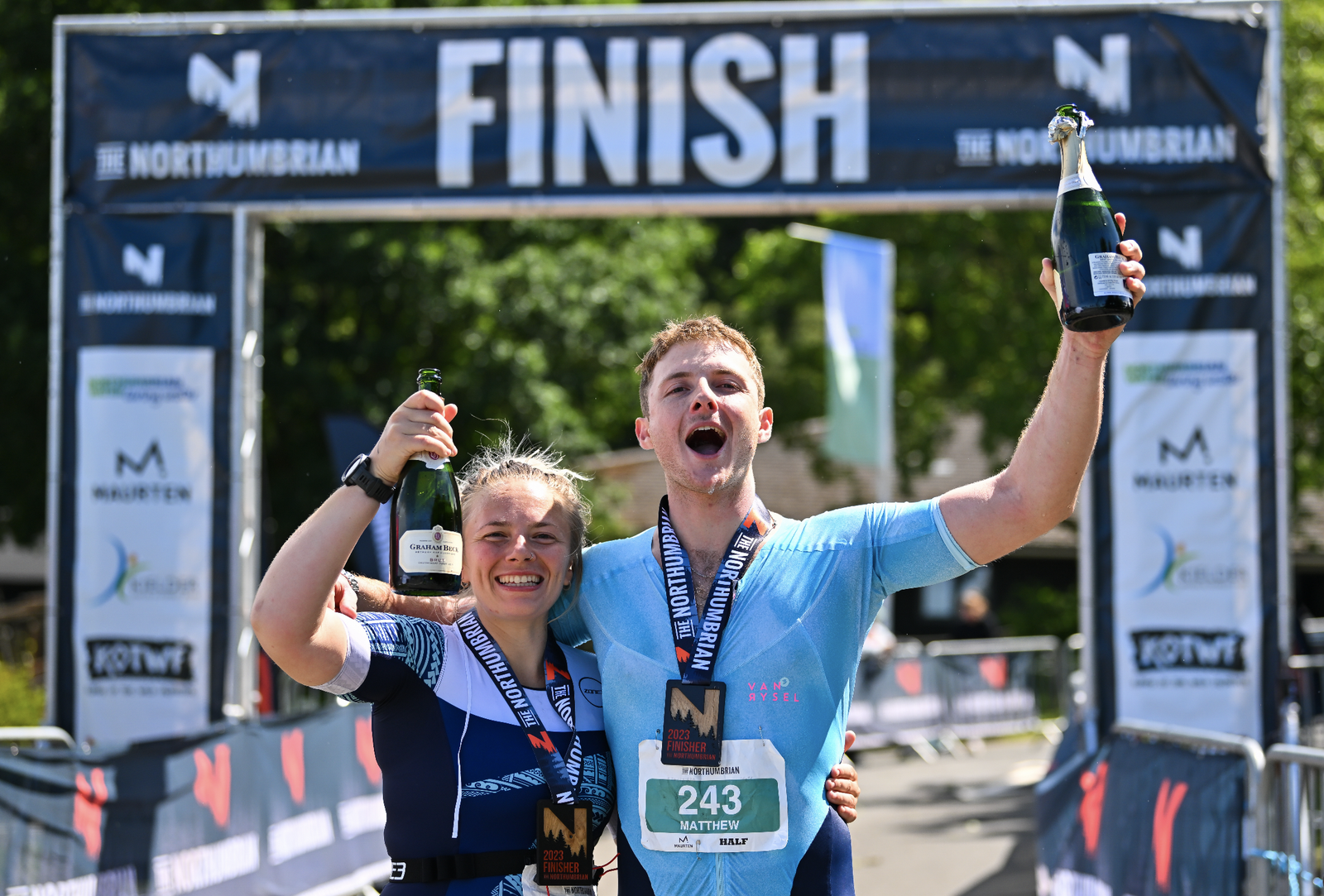Two participants celebrate at the finish line of The Northumbrian event. They are raising champagne bottles and wearing medals, smiling joyfully. The banner and crowd barriers in the background highlight the festive atmosphere of the race's conclusion.