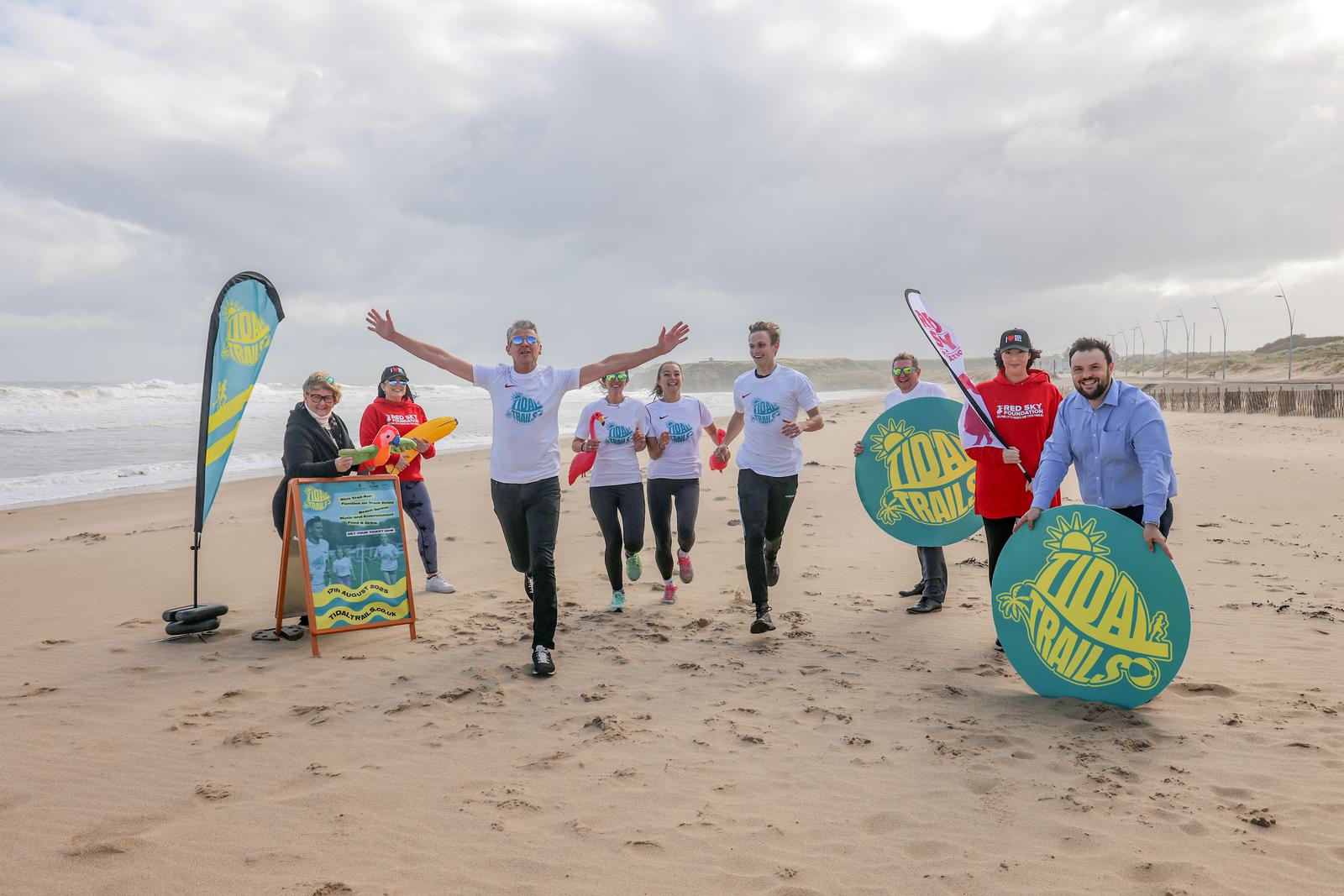 A group of people in running attire jog along a sandy beach. They hold colorful banners and a round sign with "100 Trails." The sky is cloudy, and the sea is visible in the background. A woman with a dog stands near a signboard and flag.