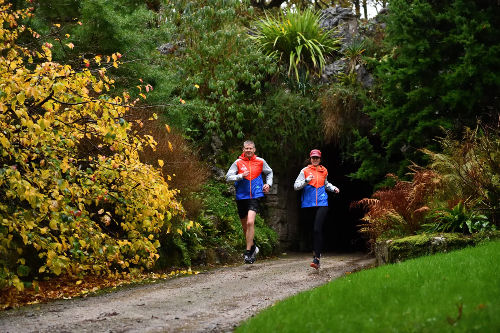 Two people running on a leafy path flanked by greenery and colorful autumn foliage. Both are wearing matching orange and blue jackets and black leggings. The path curves into a forested area, and the runners appear to be enjoying a nature trail.
