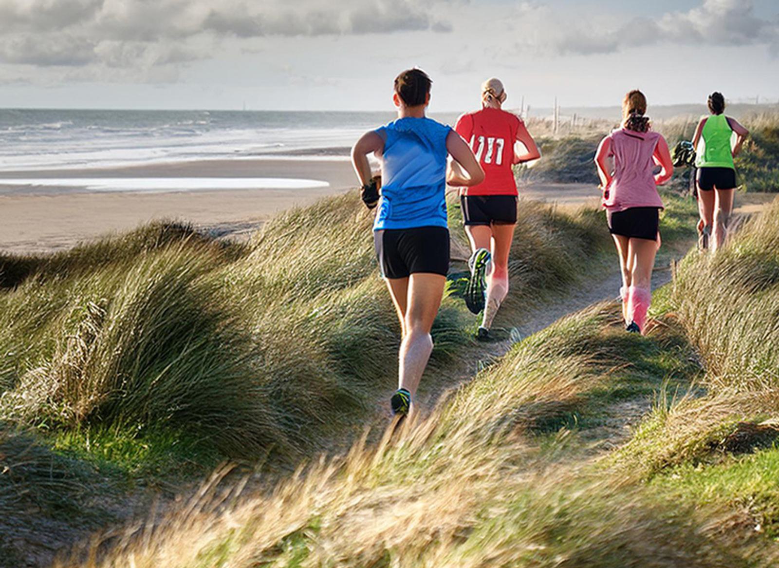 Four runners jog along a grassy path near a sandy beach under a cloudy sky. They wear athletic clothing in blue, red, pink, and green, with numbers visible on some shirts. The ocean is in the background.