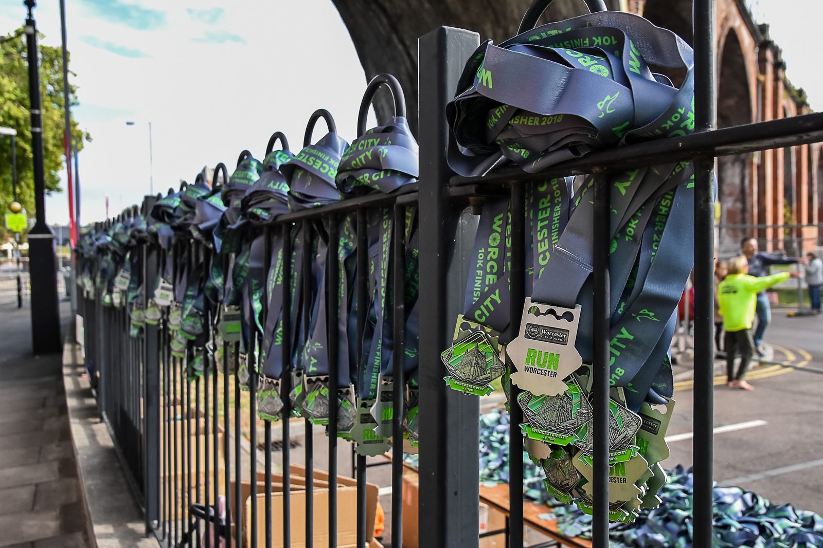 A row of medals hanging on black metal railings, each with a blue and green ribbon labeled "Worcester City Run 2017." The background shows a street scene with people and the arch of a bridge. The foreground includes medal boxes on the ground.