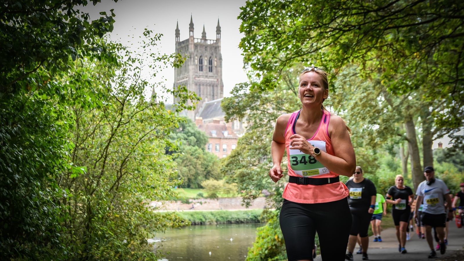 A woman wearing a pink tank top and black leggings, with race number 348, runs along a path in a park. Other runners are seen in the background, and a historic building with towers is visible beyond the trees. The scene is green and lush, indicating a summer or spring day.