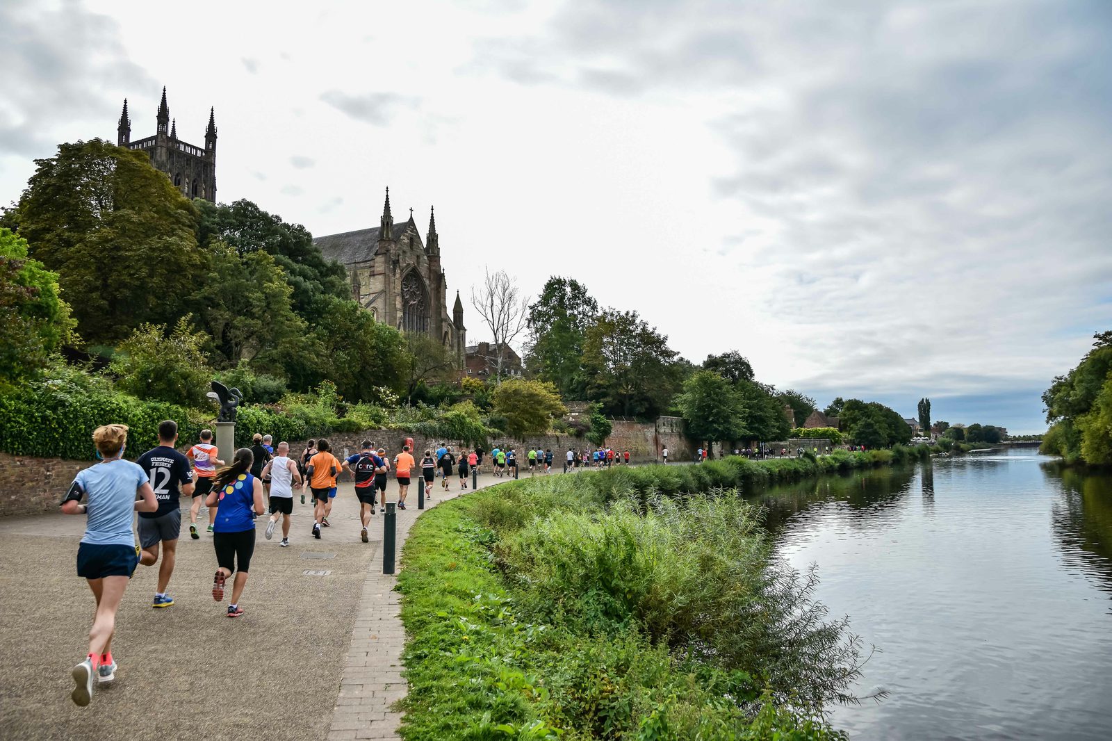 Runners participating in a race along a riverside path, with trees and greenery on one side and a body of water on the other. A historic cathedral is visible in the background to the left, set against a partly cloudy sky.