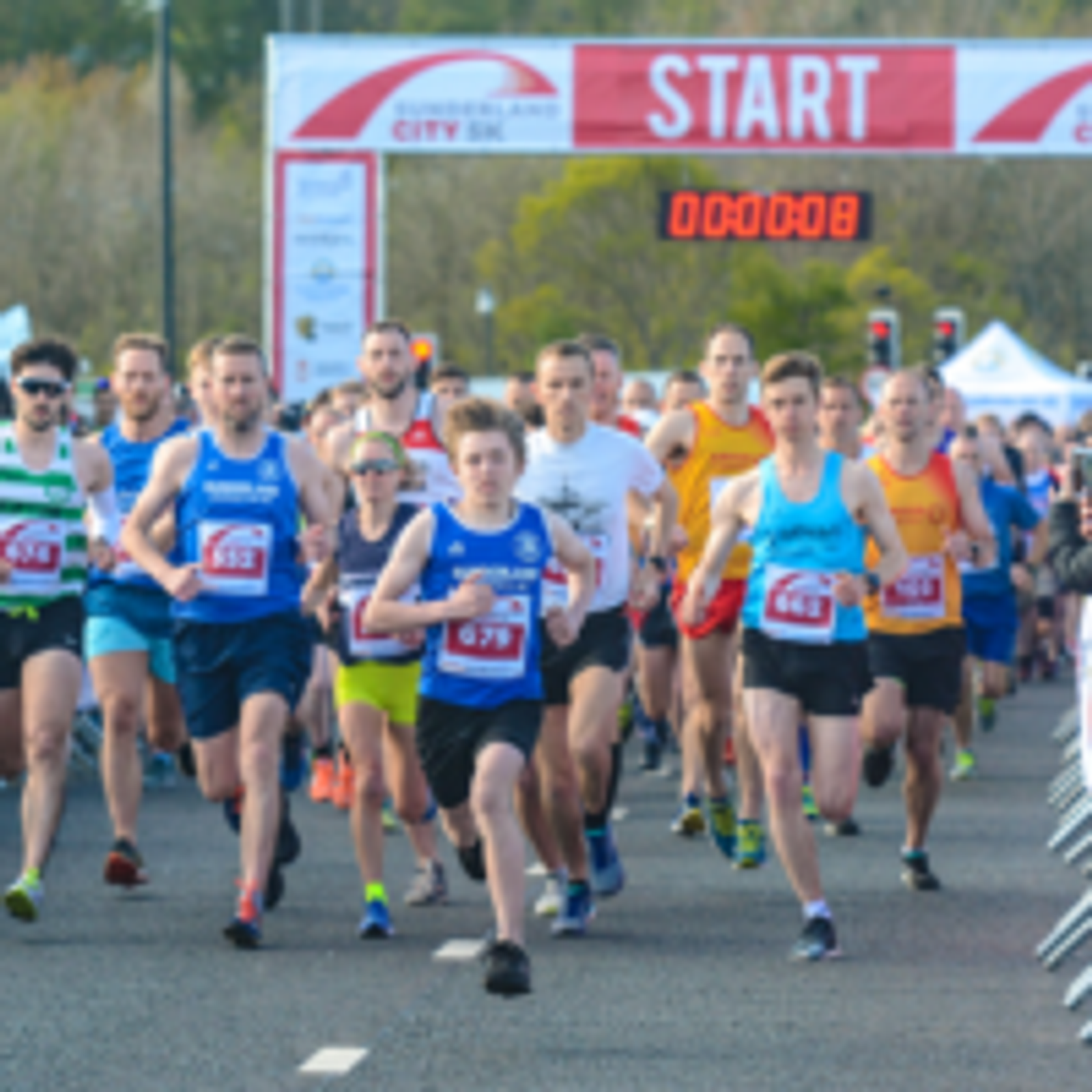 Runners in race bibs start running at a road race, with a "START" banner overhead and a crowd behind them. The digital timer above the banner shows "00:00:08." The runners wear varied athletic gear, and the background includes trees and race signage.