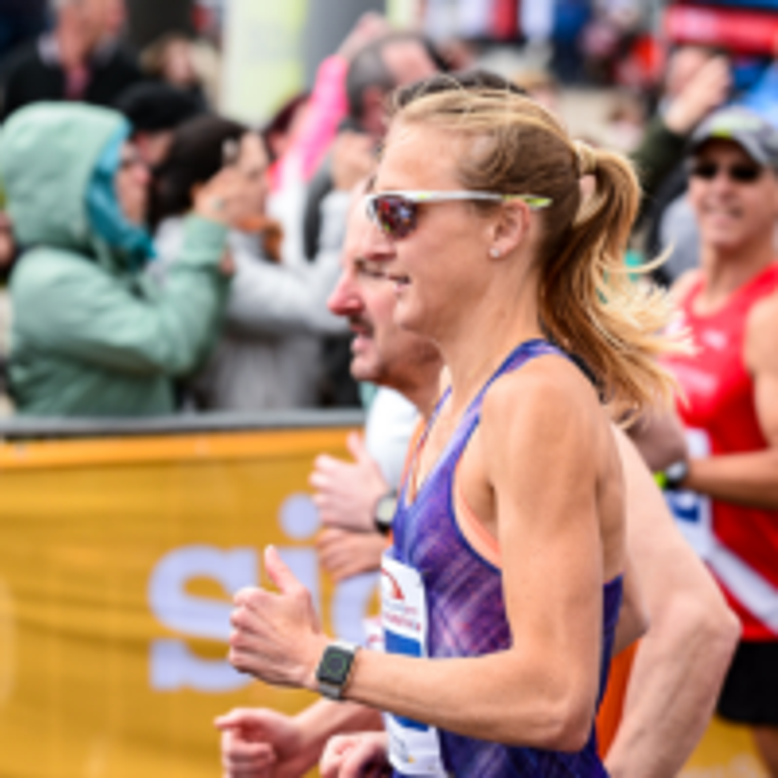 A female runner with sunglasses, a smartwatch, and a purple tank top is participating in a race. She is surrounded by other runners and spectators, some of whom are blurred in the background.