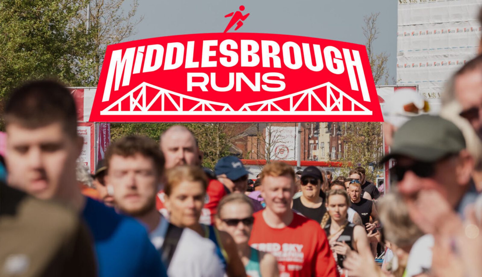 A crowd of runners outdoors with a large red and white "Middlesbrough Runs" logo featuring a bridge graphic displayed prominently above them.