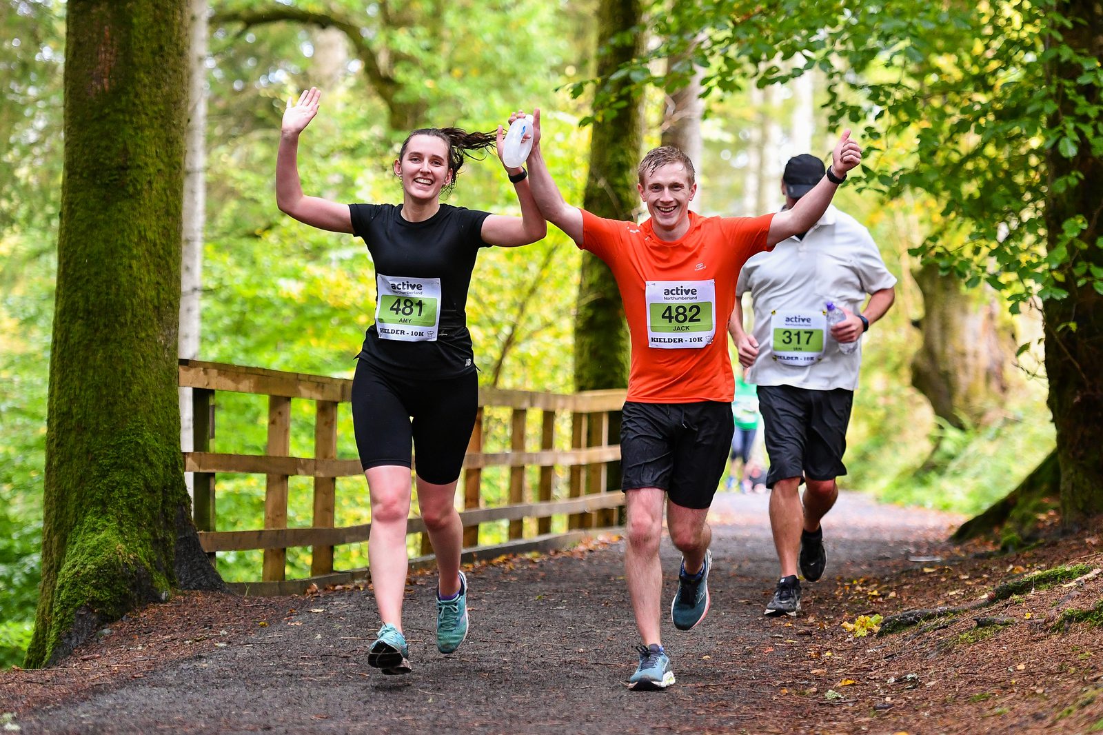 Two runners in athletic gear cheer as they hold hands and raise their arms in celebration while running on a forest trail. The woman on the left wears a black outfit with bib number 481, and the man on the right wears an orange top with bib number 482. Other runners are seen in the background.