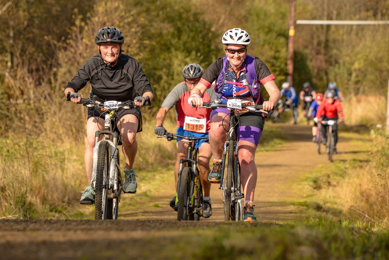 Two cyclists, a man and a woman, are riding mountain bikes on a dirt path during a race. They are smiling and wearing helmets. Other cyclists are visible behind them, with numbers on their bikes. The background is a mix of trees and an open field.