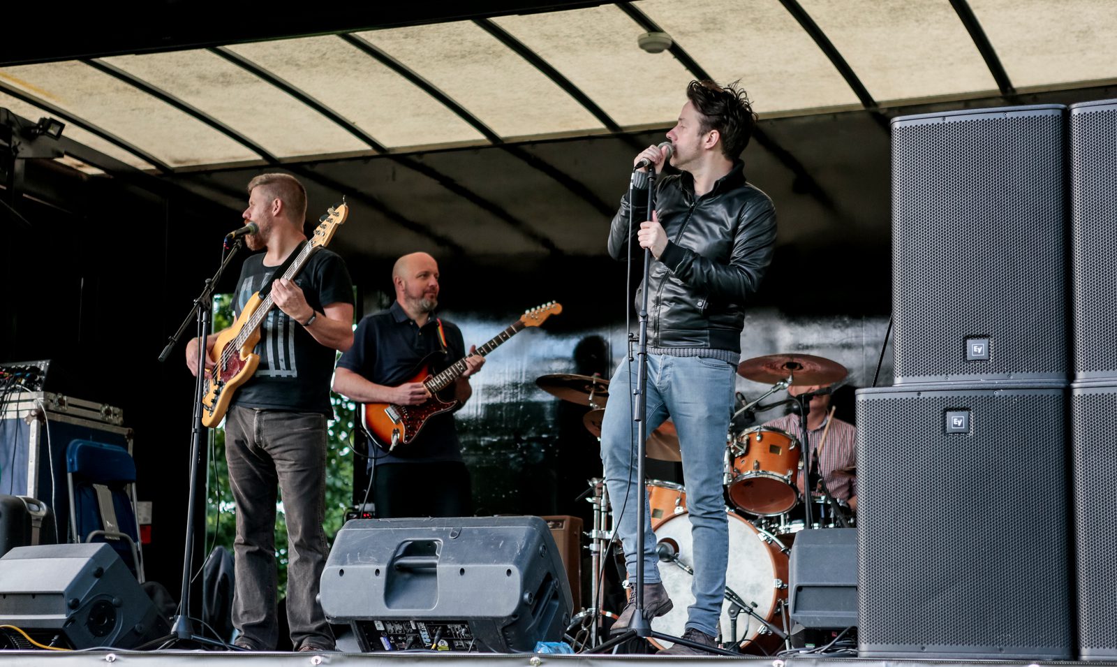 A live band performs on an outdoor stage. The group includes a bassist, a guitarist, a drummer, and a lead singer. The male lead singer is holding a microphone and wearing a black leather jacket. Sound equipment and large speakers are visible in the foreground.