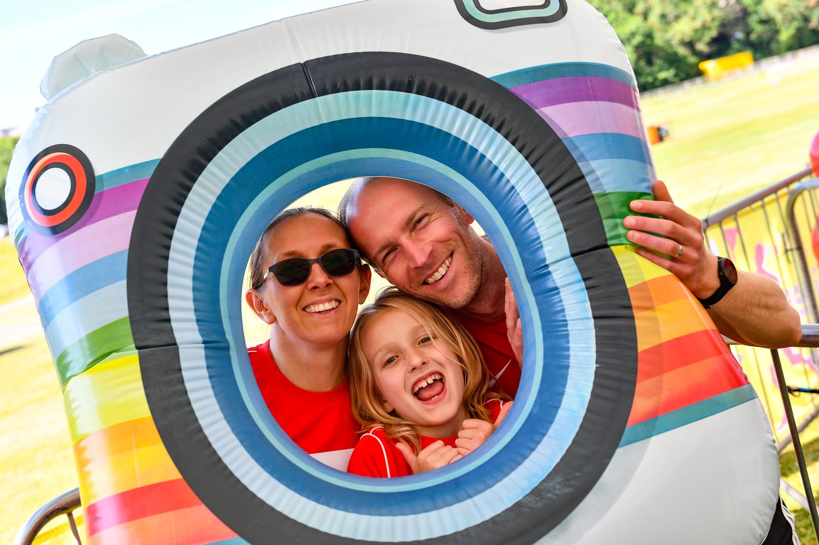 A happy family of three pose while smiling through the circular opening of a colorful, inflatable photo frame. They are outdoors, on a sunny day, with green grass and trees in the background. The family members are wearing red shirts and appear joyful and excited.