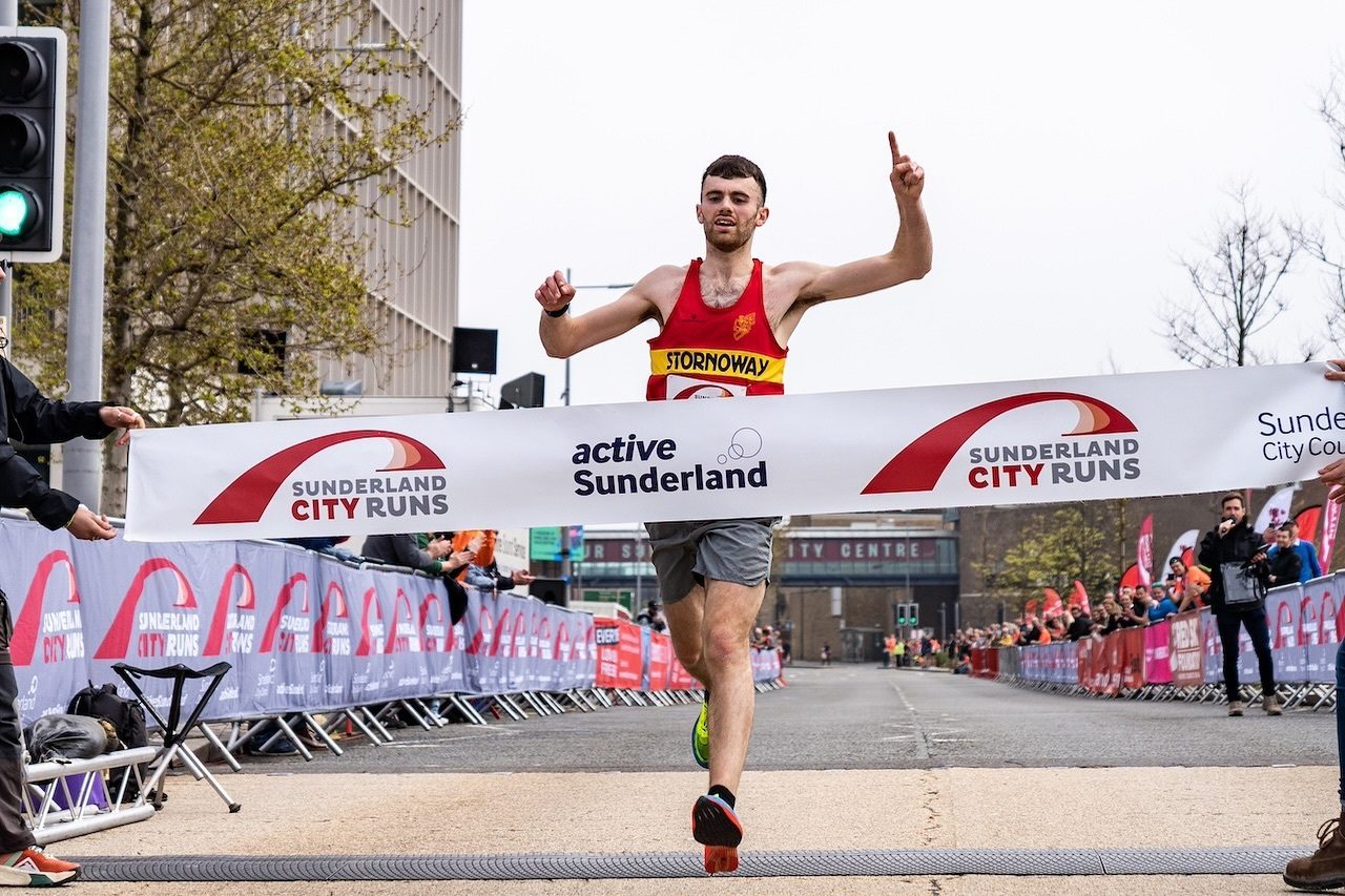 A male runner, wearing a red and yellow tank top labeled "STORNOWAY," crosses the finish line at a Sunderland City Run event, raising one finger in victory. The banner reads "active Sunderland" and "Sunderland City Runs." Spectators and other participants are in the background.