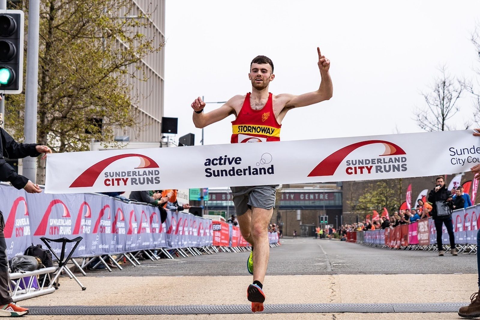 A male runner, wearing a red and yellow tank top labeled "STORNOWAY," crosses the finish line at a Sunderland City Run event, raising one finger in victory. The banner reads "active Sunderland" and "Sunderland City Runs." Spectators and other participants are in the background.