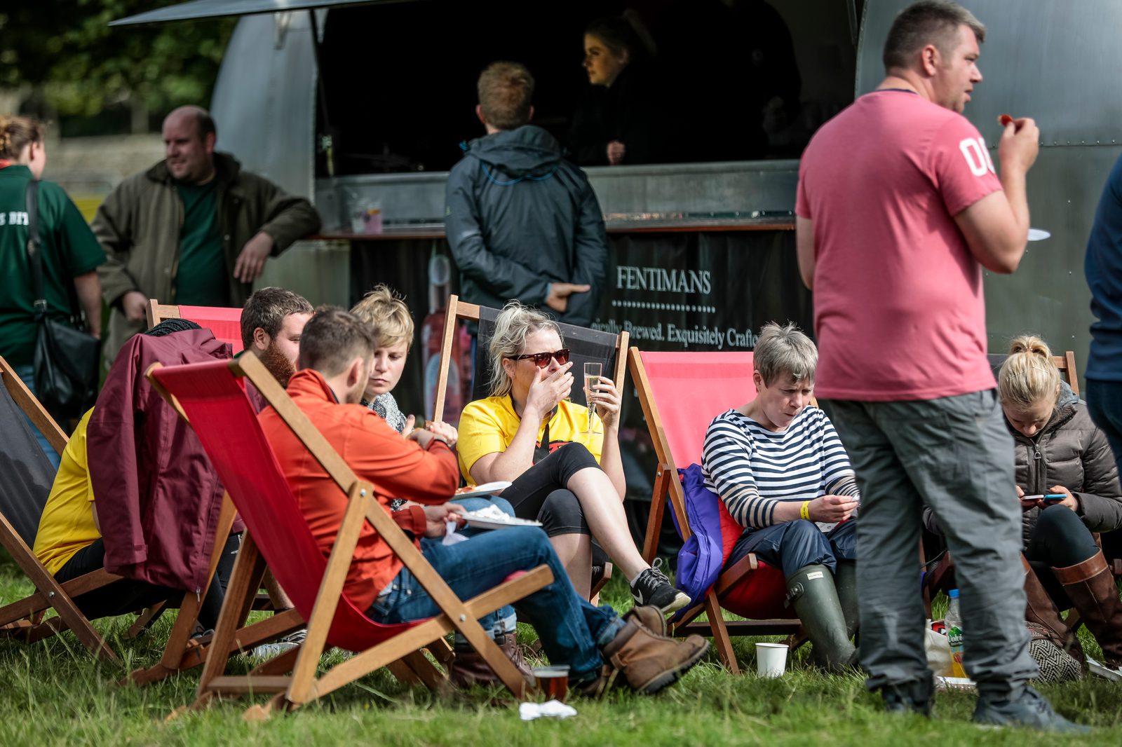 A group of people, including men and women, sit on deck chairs in front of a food truck with a "Fentimans" sign. Some are eating, others are on their phones, and a few are chatting. In the background, more people are gathered near the truck. The scene is a casual outdoor setting.