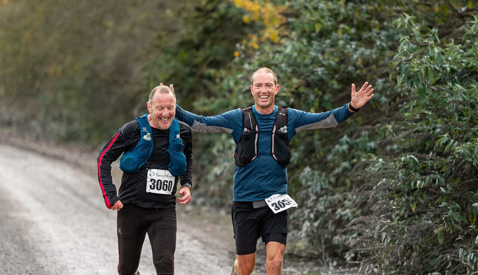 Two men running a trail race with smiles, wearing race bibs 3060 and 3059. They are on a dirt path, surrounded by lush greenery. The event logo "Andover Trail Events" is visible in the corner.