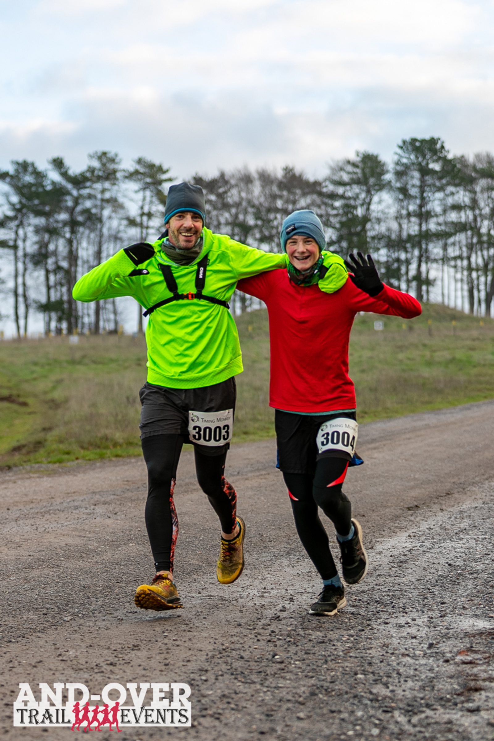 Two runners wearing bright gear and beanies, smiling with arms around each other, participate in a trail event. They have bib numbers 3003 and 3004. A trail and trees are visible in the background under a cloudy sky. "Andover Trail Events" logo at the bottom.