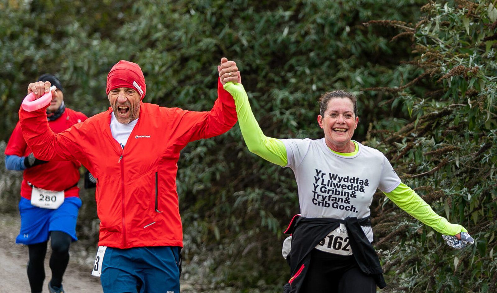 Two runners, a man in a red jacket and a woman in a white shirt, cheerfully holding hands as they cross a finish line in a trail event. They appear joyful and triumphant, with greenery in the background.