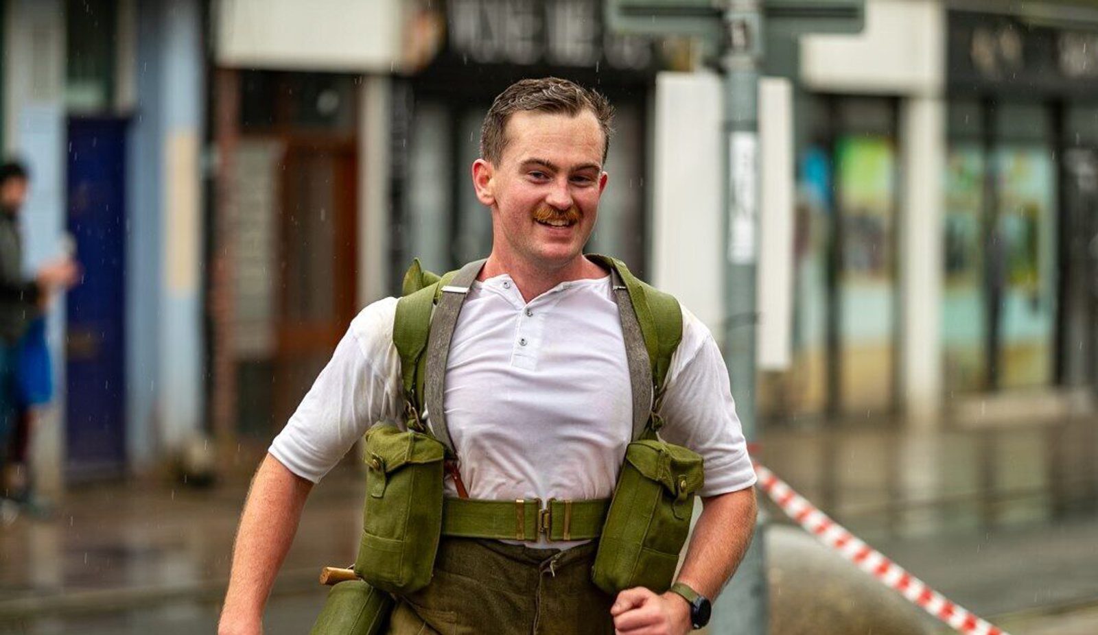 A person runs outdoors carrying heavy gear, appearing determined and focused. They're wearing a white shirt and olive-colored gear. The background shows an urban setting with rain, blurred shops, and a caution tape off to the side.