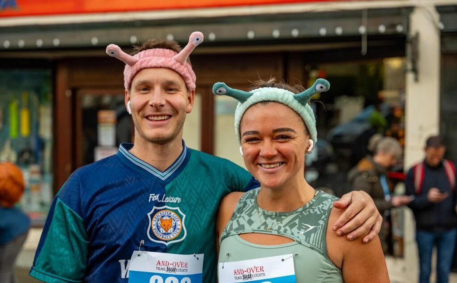 Two runners in colorful snail hats and sporting bib numbers 202 and 203 smile in front of a building during an And-Over Trail Events race. People in the background can be seen milling about.