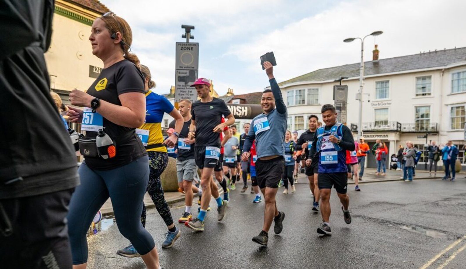 Runners participating in a marathon, wearing race numbers and athletic gear, pass through a town street. One runner raises a hand with a phone. Buildings and spectators are in the background. Event branding visible in the corner.