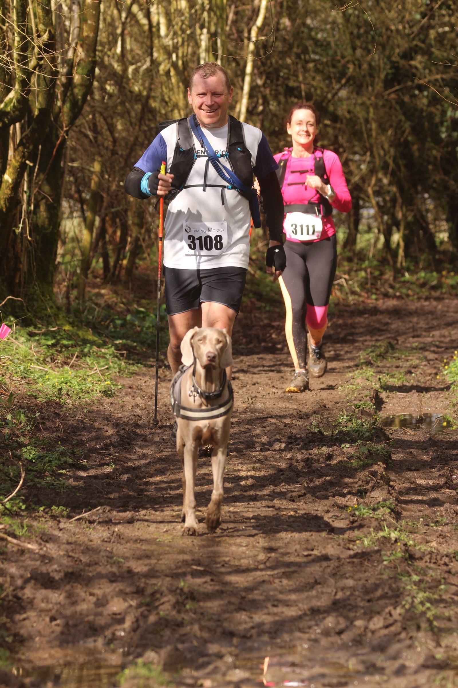 Two runners with numbered bibs run on a muddy trail through a forest. A dog leads them on a leash. The man in a white shirt and the woman in a pink top both appear joyful as they enjoy the outdoor race.